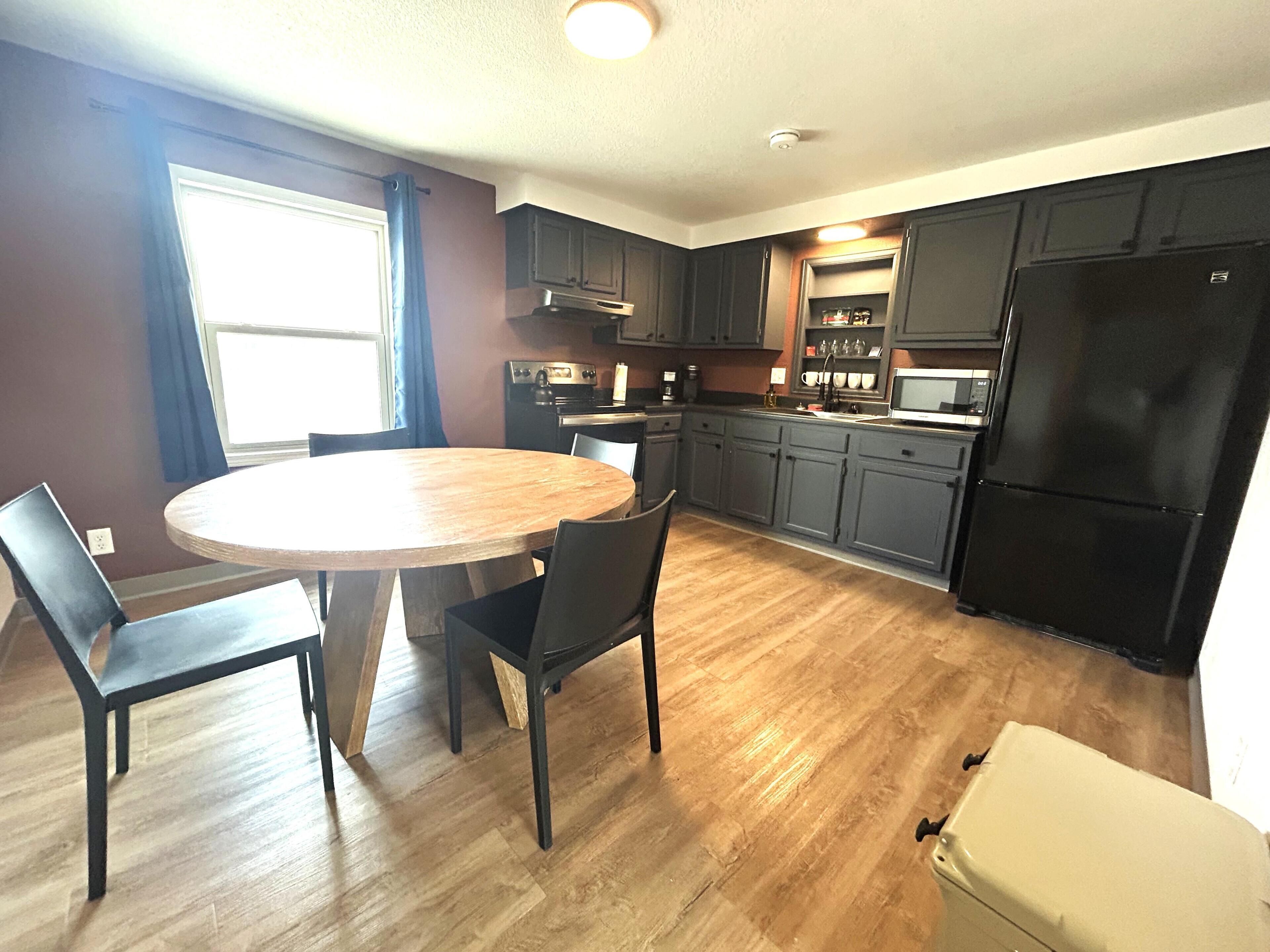 A kitchen with a dining table, chairs, cabinets, a refrigerator, and a window.