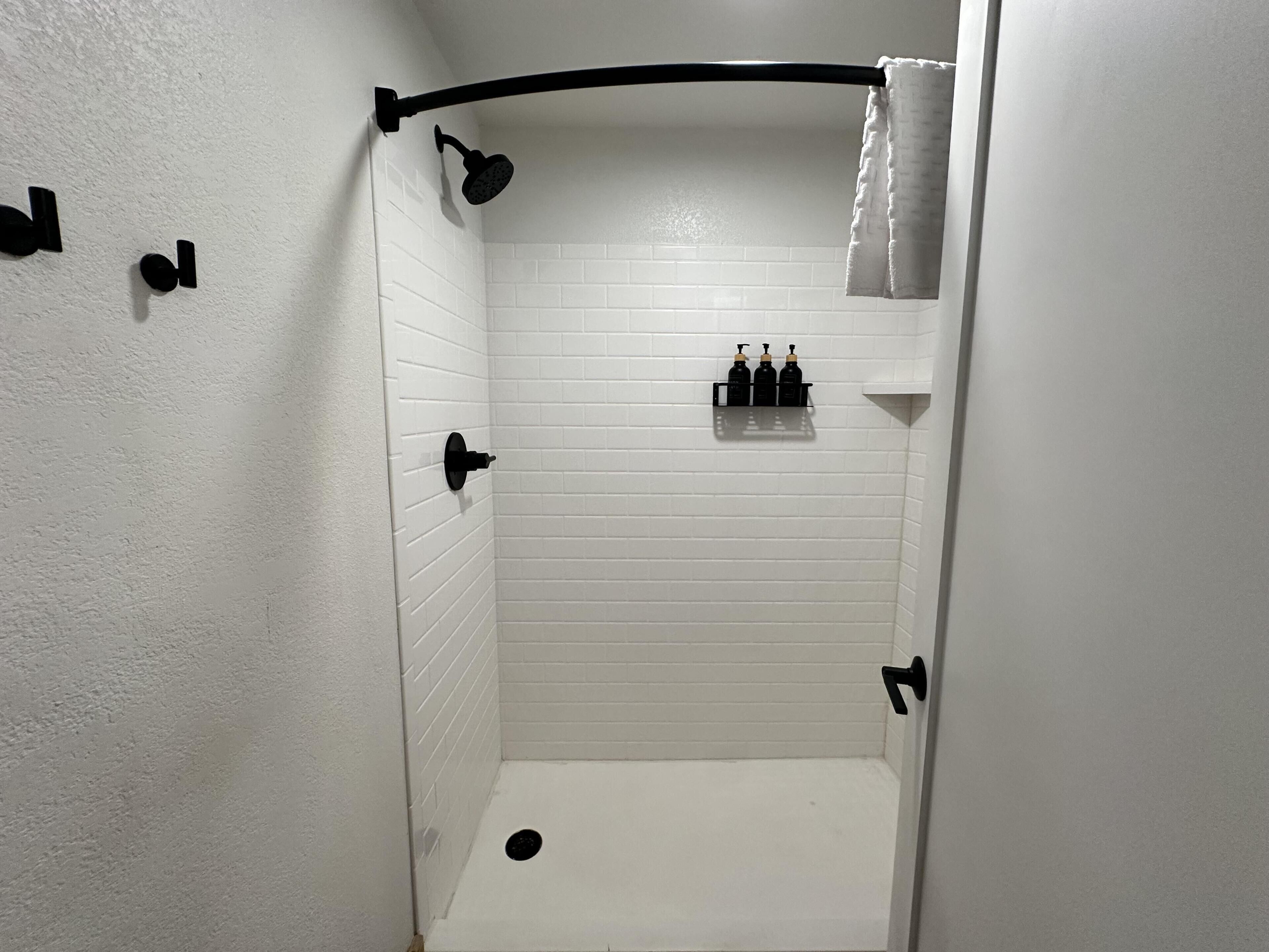 Un pequeño box de ducha con azulejos blancos, un cabezal de ducha negro y un perchero para toallas.