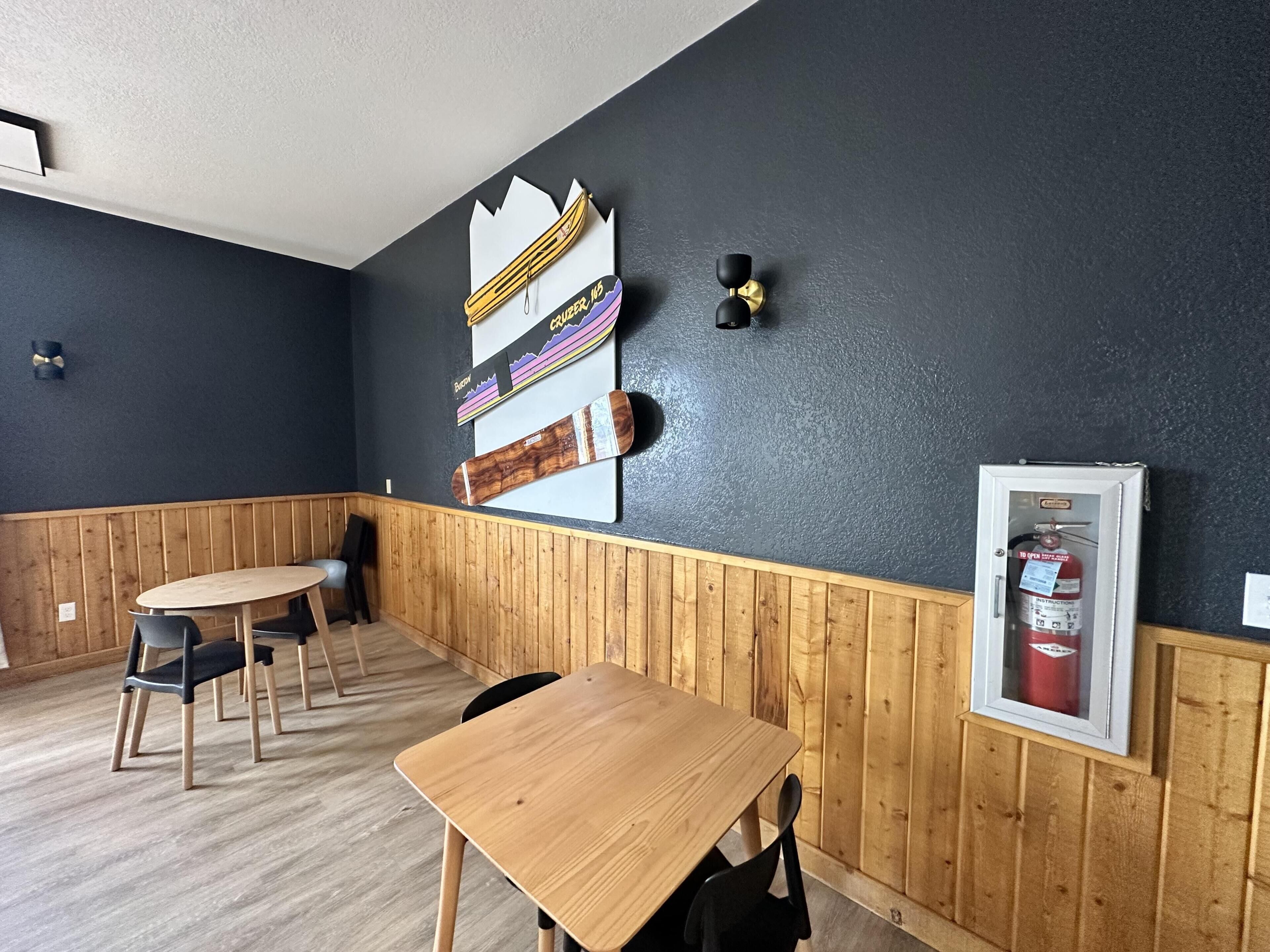 Una habitación con una mesa y sillas de madera, una pared negra decorada con una tabla de snowboard y esquíes, y un extintor.