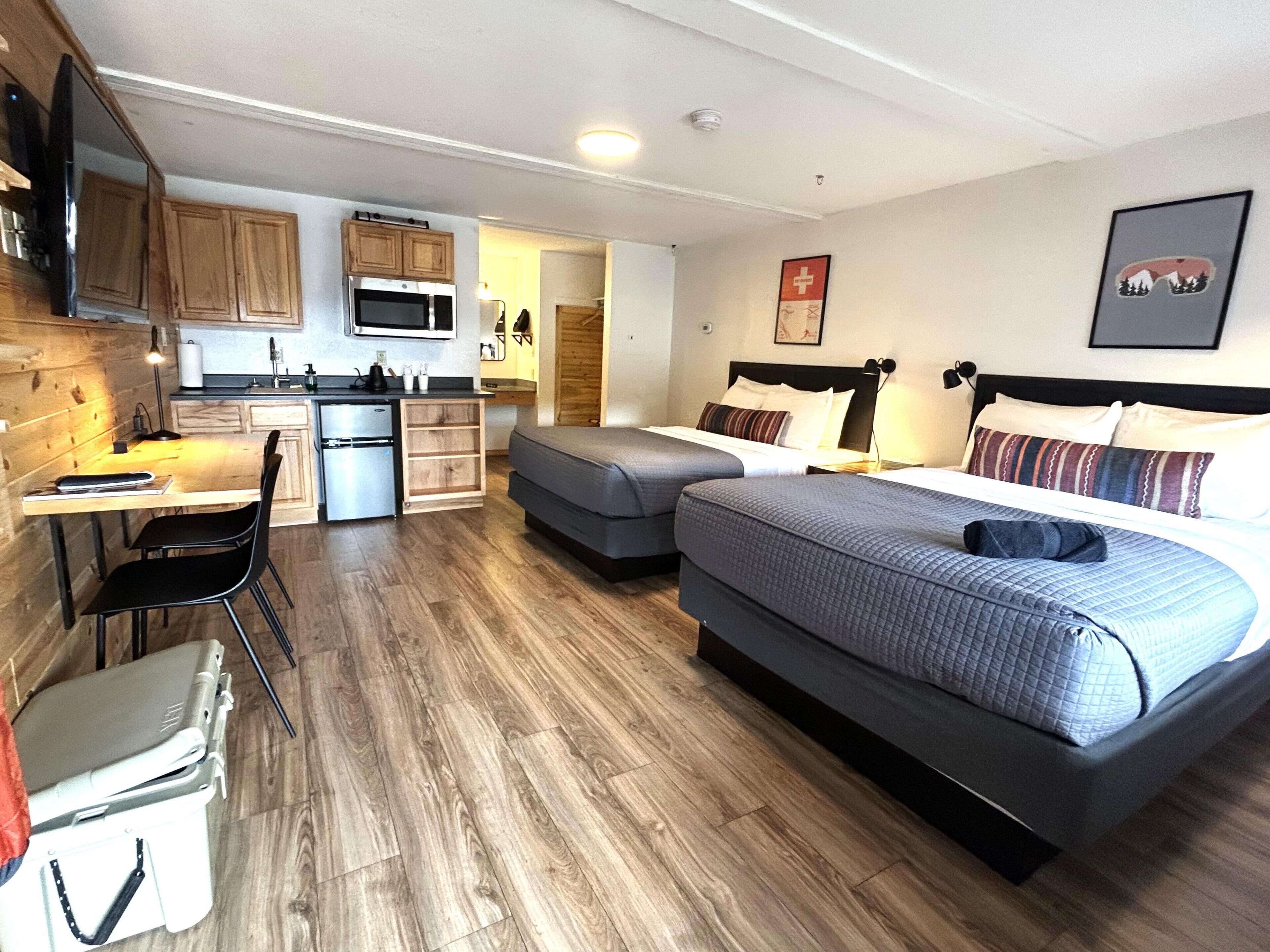 A compact hotel room with a kitchenette, a bed with two pillows, a desk with a chair, and a small storage unit.