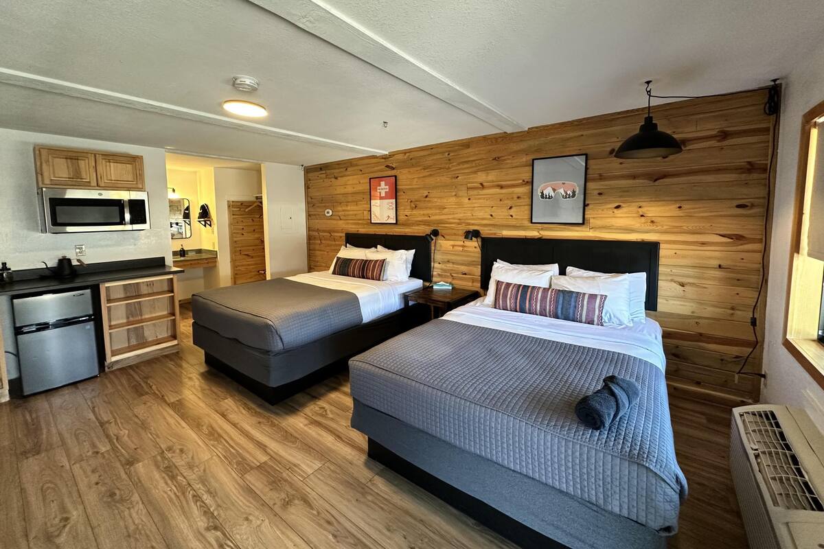 A room with two beds, a kitchenette, wooden walls, and a ceiling light.