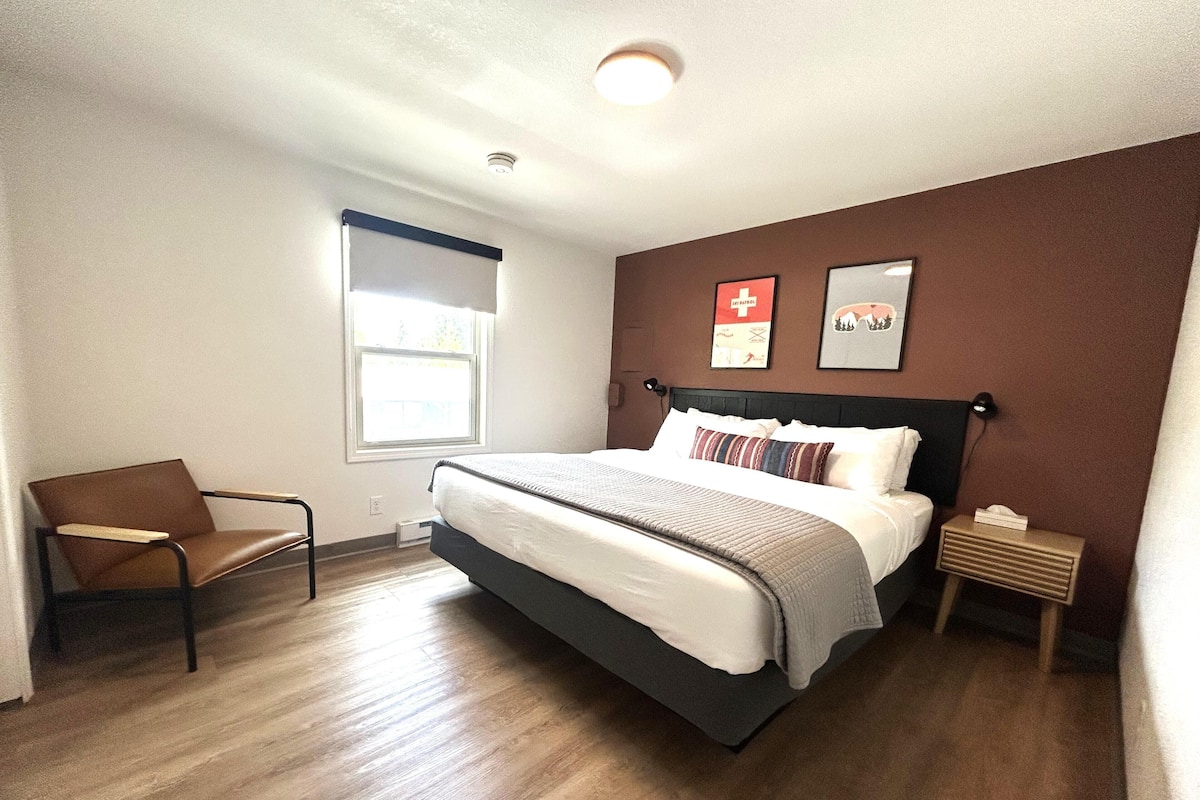 A bedroom with a bed, a chair, a nightstand, a window, and two wall-mounted pictures.