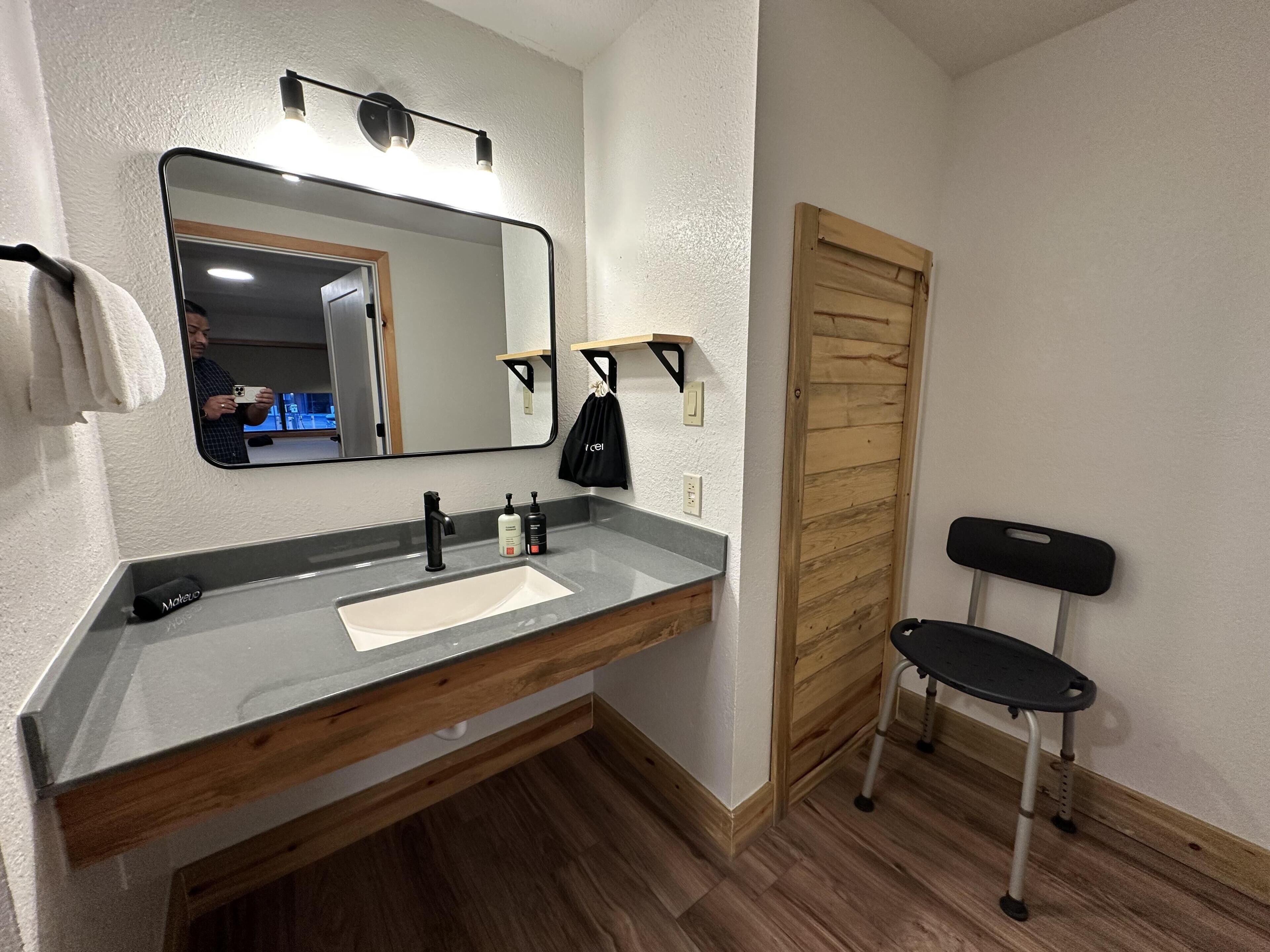 A bathroom with a large mirror, a vanity with a sink, a towel rack, a wooden door, and a wheelchair.