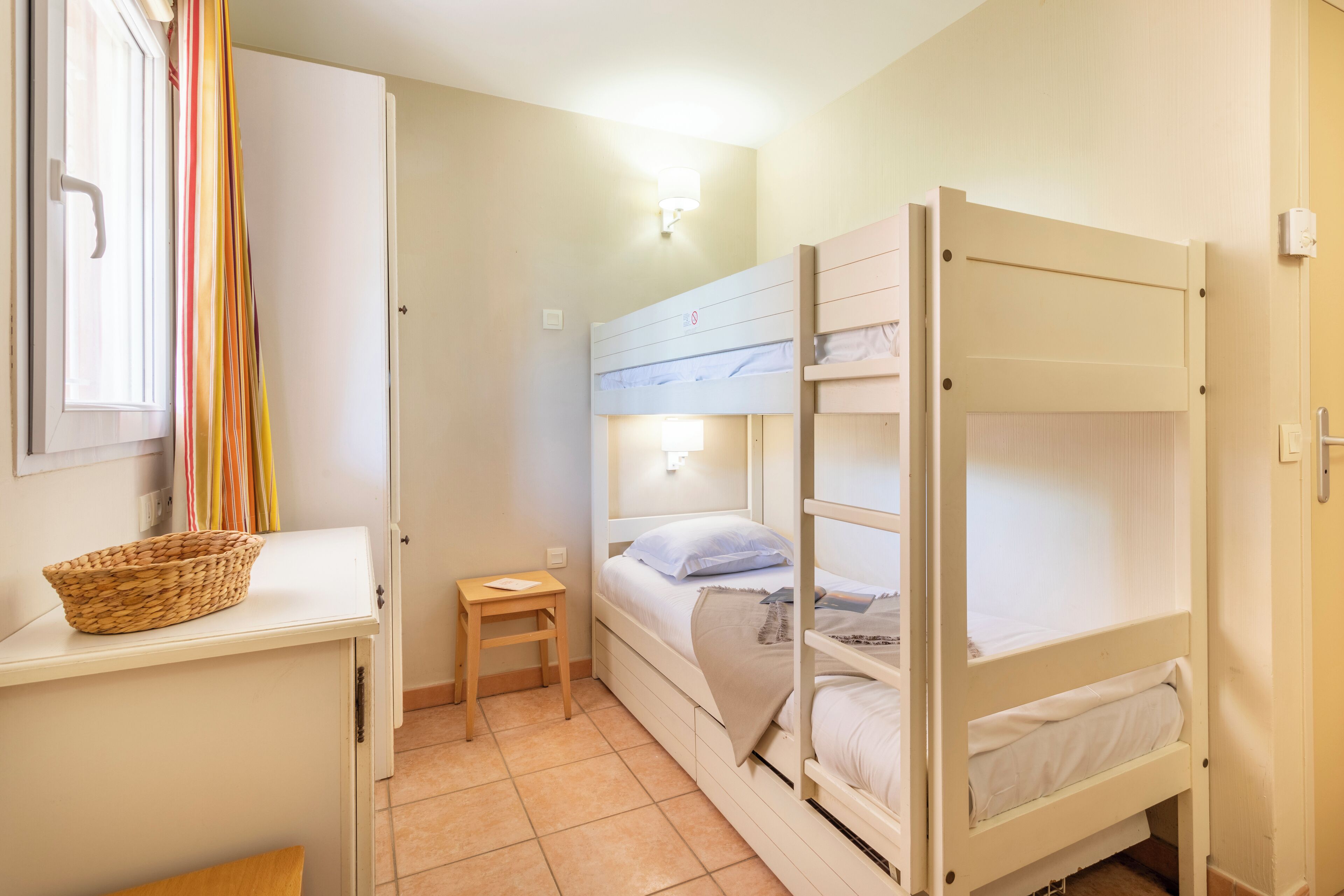Une chambre de dortoir avec des lits superposés, un petit bureau avec un panier, une table de chevet et une fenêtre avec des rideaux.