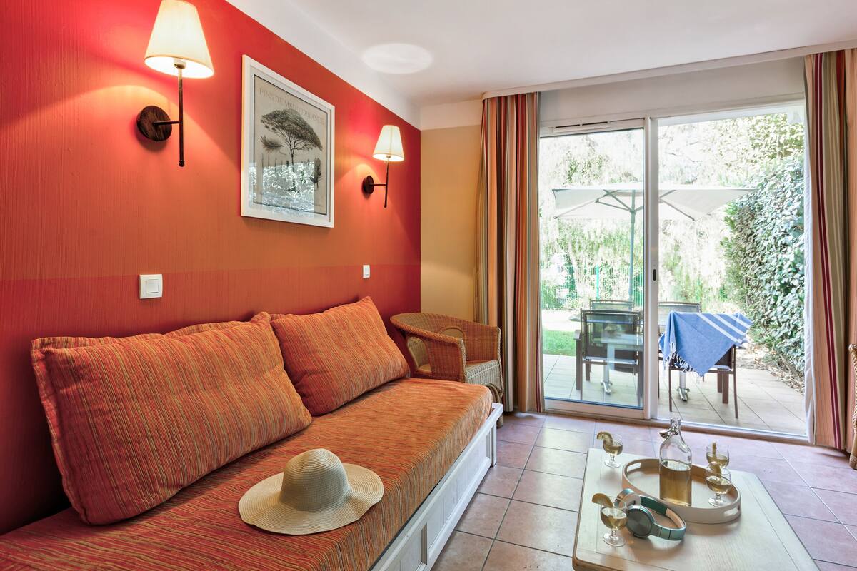 A room with a red wall, a sofa with striped cushions, a hat on the sofa, and a table with a tray of glasses and a bottle.