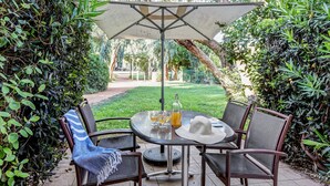 Een terras buiten met een ronde tafel, vier stoelen, een grote parasol en een fles drank.