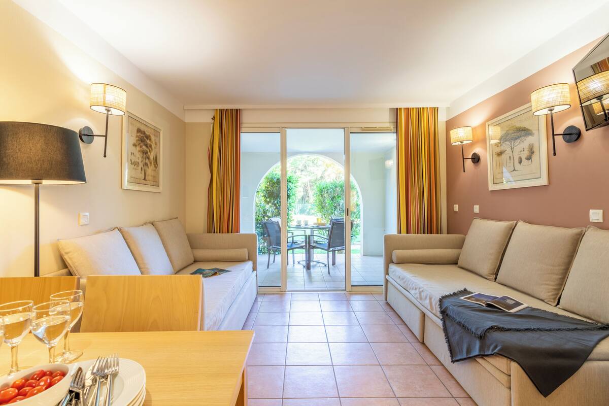 A modern living room with a sofa, a dining table with glasses and cutlery, and a view through a glass door to an outdoor area.