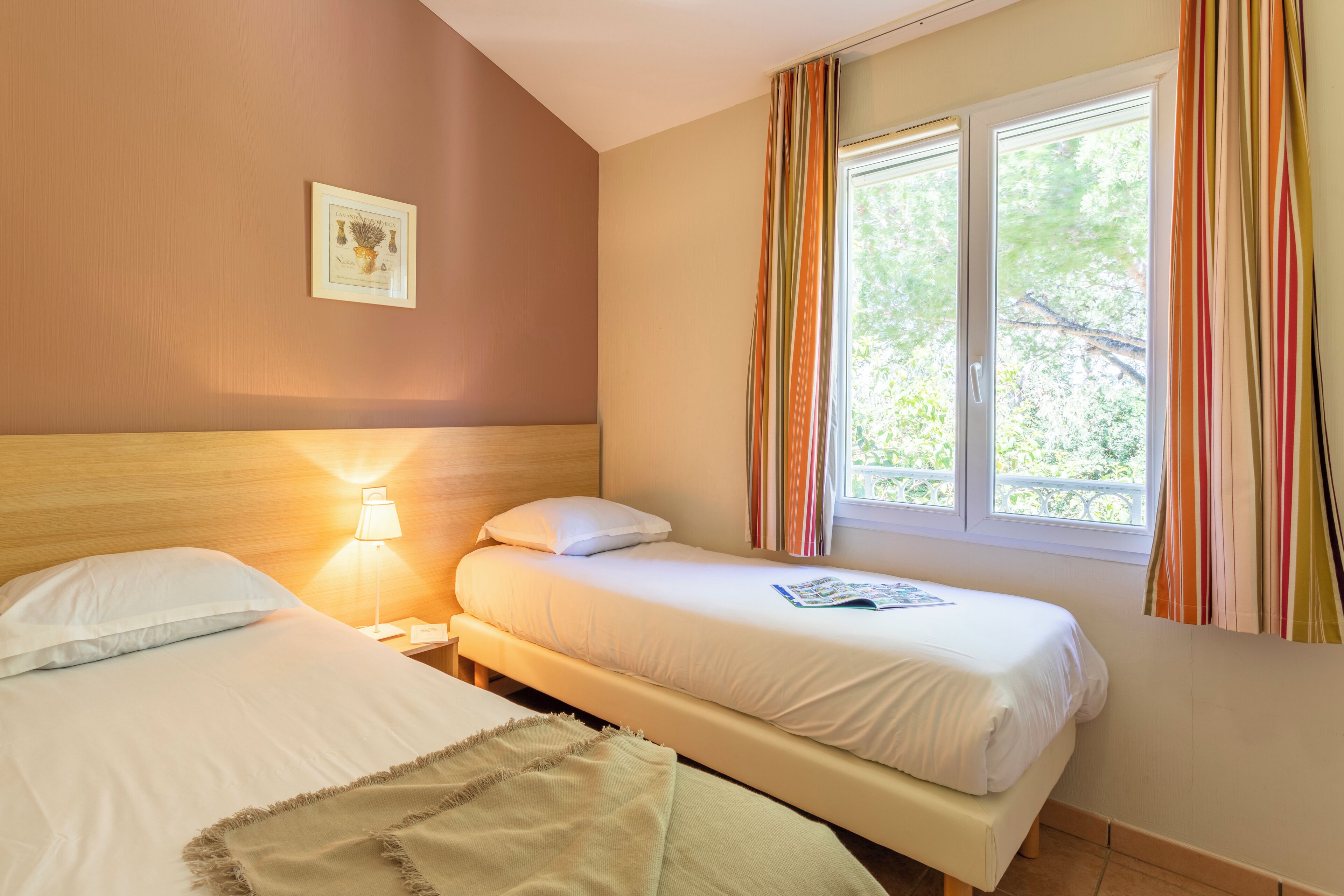 Hotellihuone, jossa on kaksi sänkyä, yöpöytä lampun kanssa, ikkuna verhoilla ja seinällä oleva kehystetty kuva.