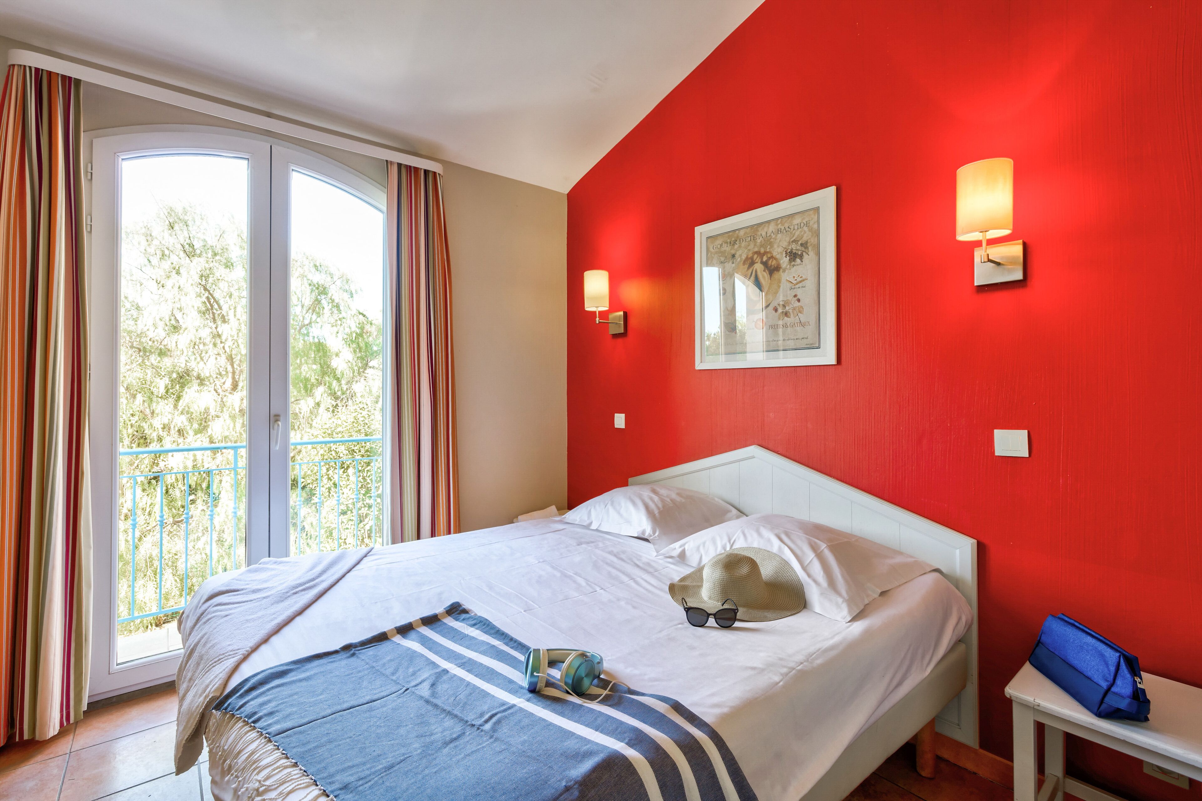 A hotel room with a bed, a window with curtains, a wall-mounted lamp, and a small table with a bag.