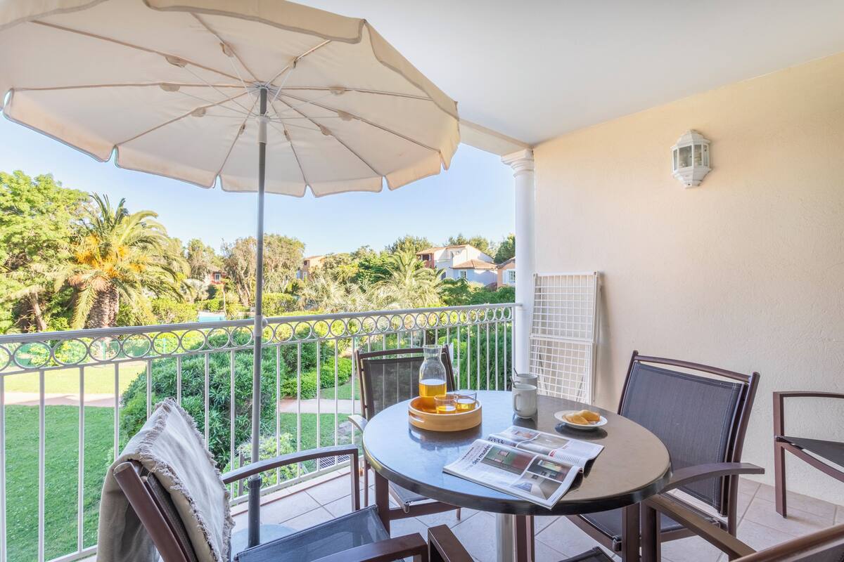 A balcony with a table, chairs, a white umbrella, and a view of greenery and houses.