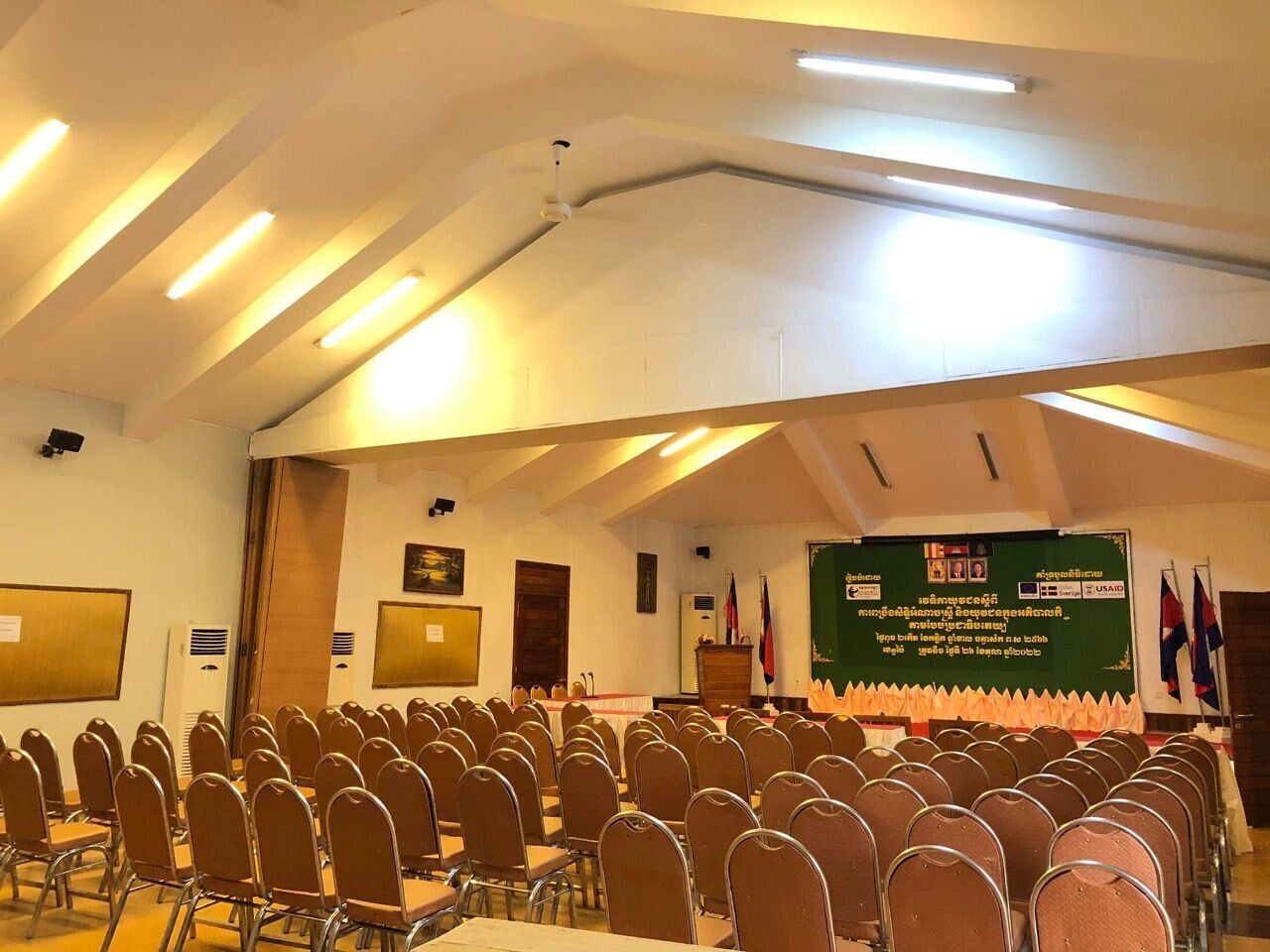 Un salón de conferencias con filas de sillas, un atril y un escenario con banderas.