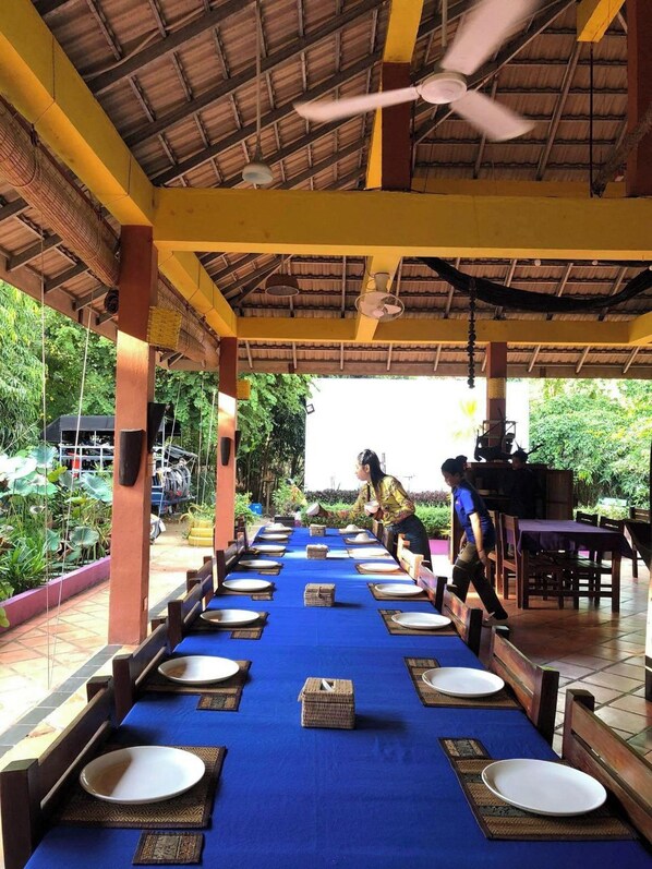 A long dining table with blue tablecloth, white plates, and woven placemats, set up outdoors with a thatched roof and wooden pillars.