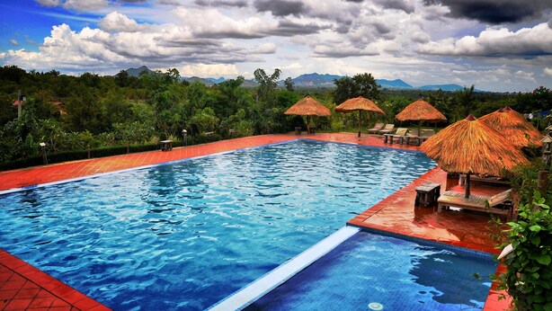 A swimming pool with a red border, surrounded by lounge chairs and thatched umbrellas, with mountains in the background. - Memoria Palace & Resort (Pailin)