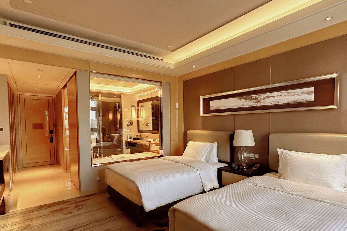 A hotel room with two beds, a large mirror, a bedside table with a lamp, and a painting on the wall.