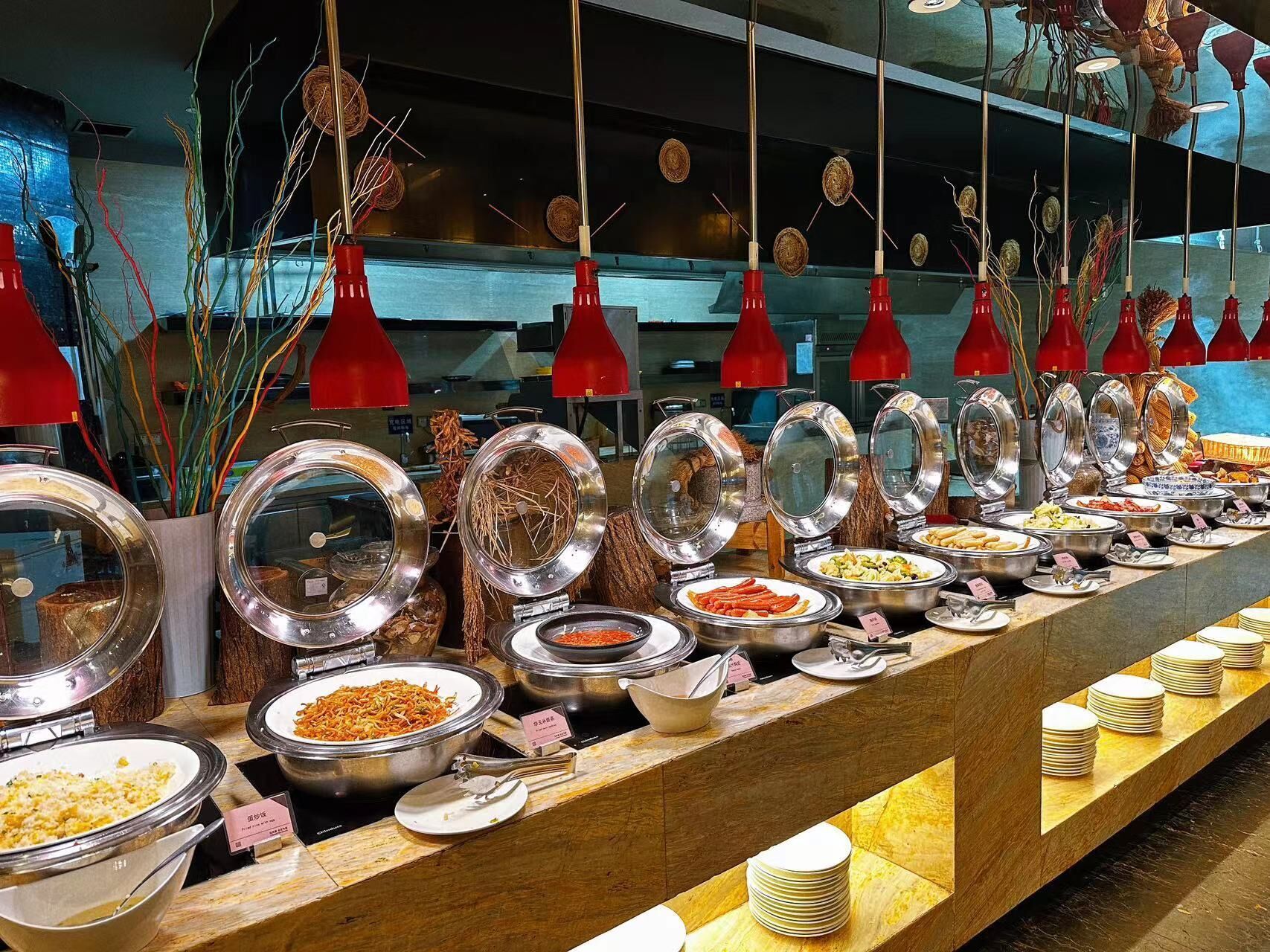 Ein Buffet mit verschiedenen Speisen in Metallbehältern, einige mit Deckeln, und rote Pendelleuchten darüber.