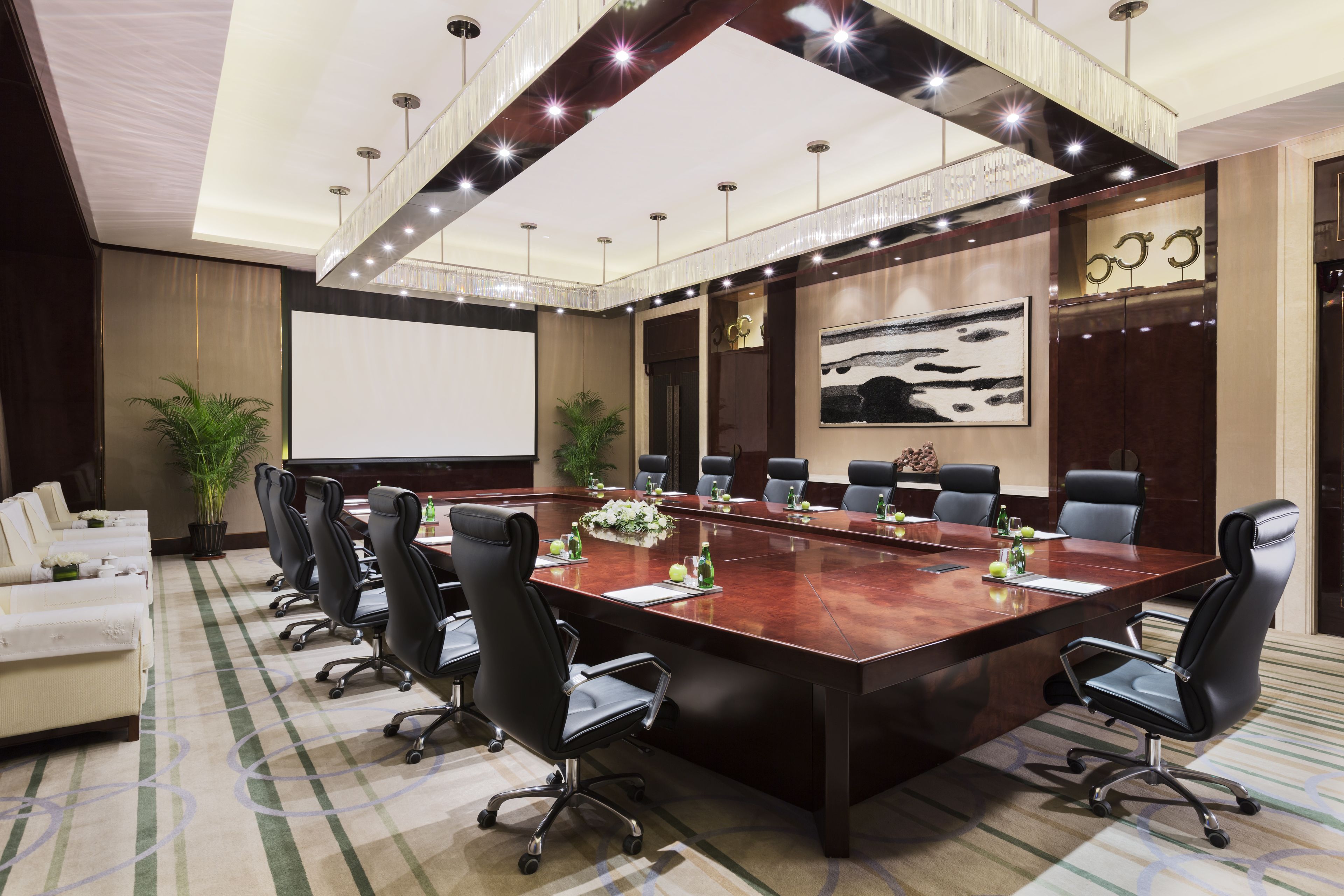 A conference room with a large oval table, black office chairs, a projector screen, and a large painting on the wall.