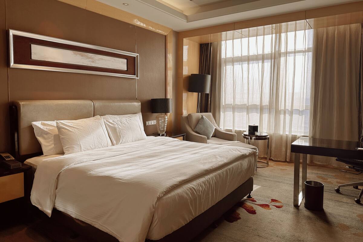 A hotel room with a large bed, a chair, a desk, and a window with sheer curtains.
