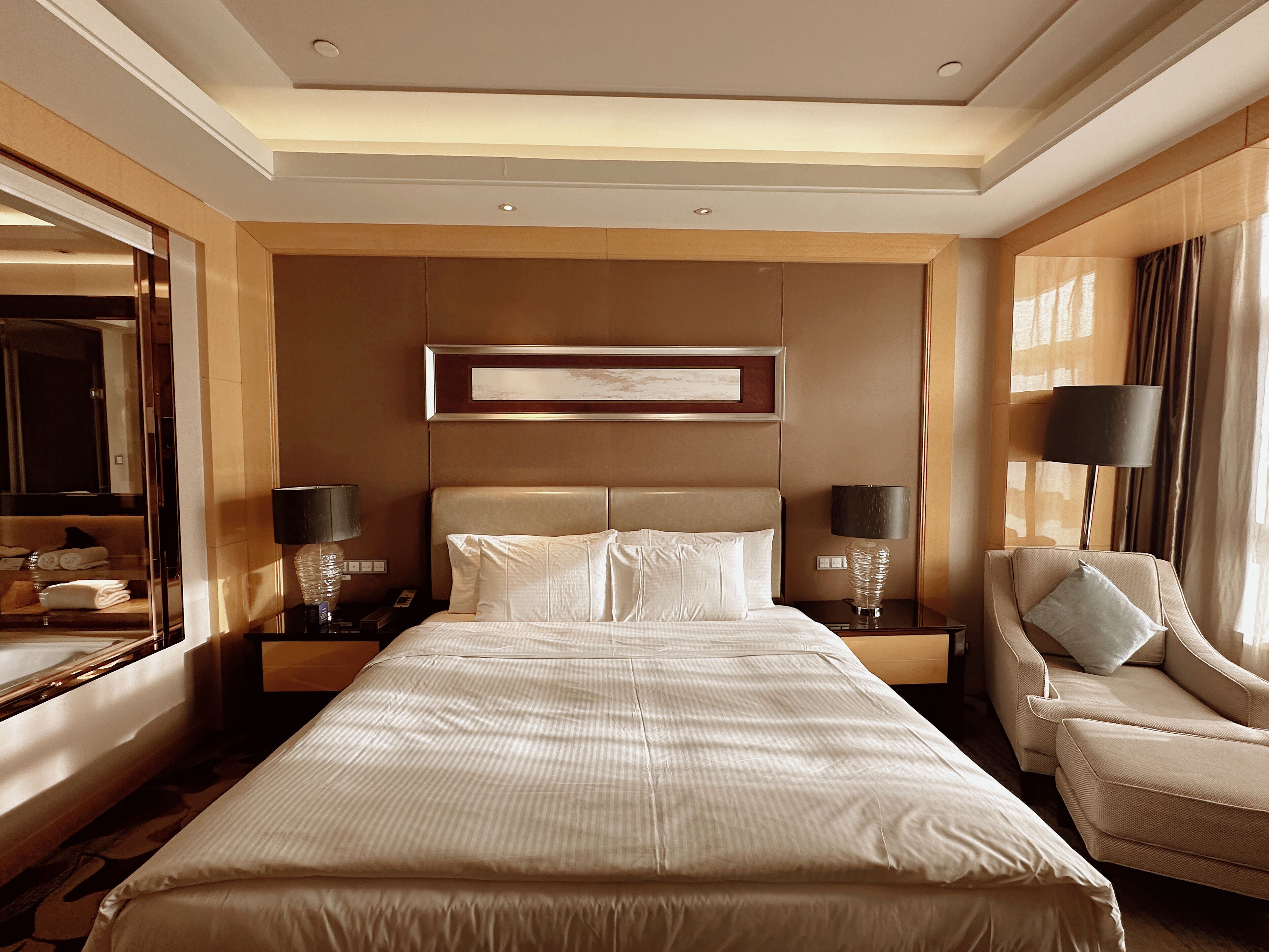 Habitación de hotel con una cama grande, dos mesitas de noche con lámparas, una silla, un espejo amplio y una obra de arte enmarcada en la pared.