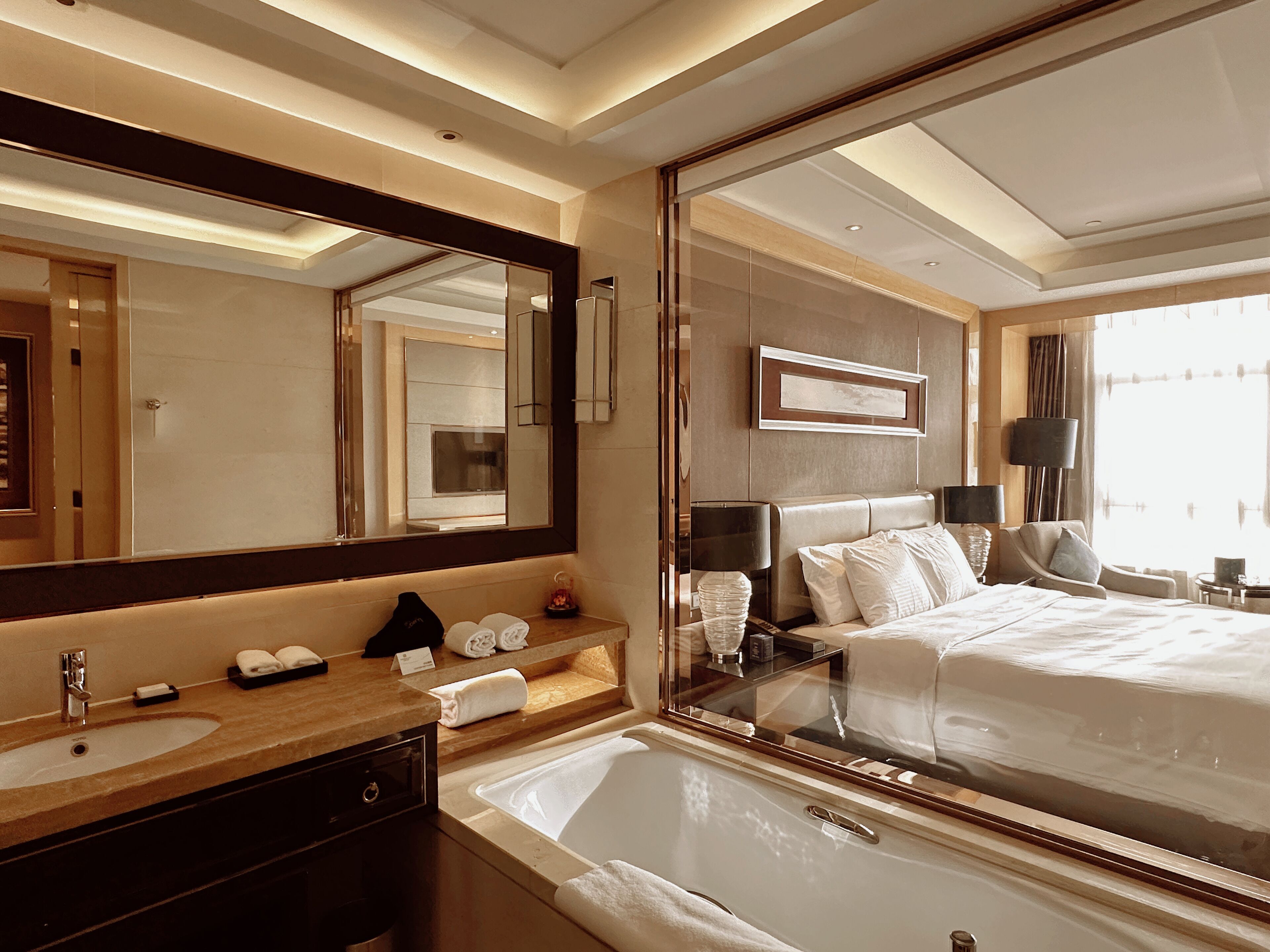Una camera d'hotel con un grande specchio, una vasca da bagno, un letto, un mobile toilette con lavandino e una finestra con tende.