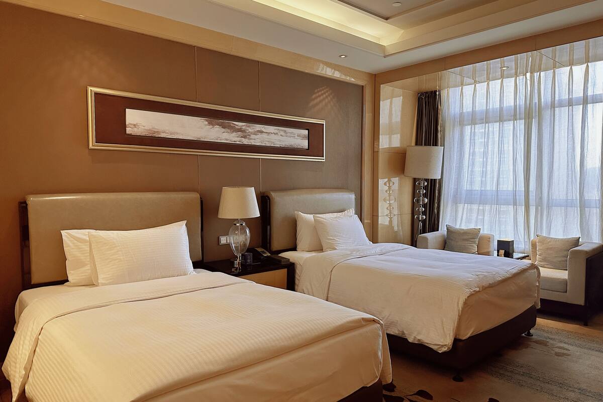 A hotel room with two beds, a large window with sheer curtains, a framed artwork above the beds, and a bedside table with a lamp.