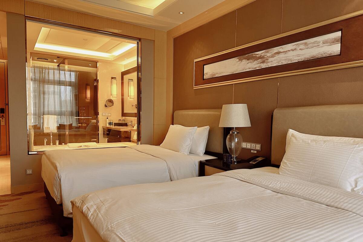 A hotel room with two beds, a nightstand with a lamp, a large mirror, and a painting on the wall.