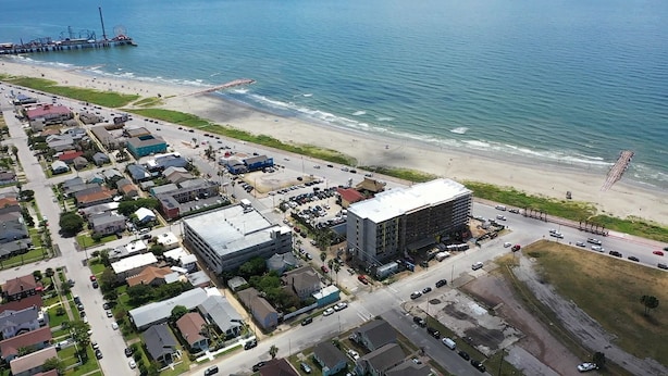 An aerial view of a coastal area with a beach, a pier, and a large building with a flat roof.
