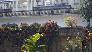 Ein Innenhof mit einem weißen Gebäude, das Bögen und ein Ziegeldach aufweist. Korbstühle und ein Tisch mit einer Kanne und Tasse sind zu sehen. Rote Blumen und Grünpflanzen sind ebenfalls sichtbar.