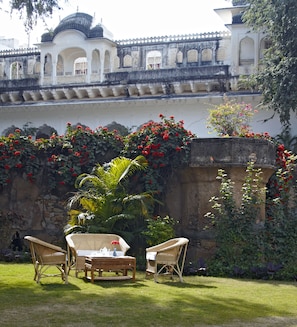 A courtyard with a white building featuring arches and a tiled roof. There are wicker chairs and a table with a pitcher and cup. Red flowers and green foliage are visible. - Samode Bagh (Chomu)