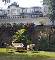 A courtyard with a white building featuring arches and a tiled roof. There are wicker chairs and a table with a pitcher and cup. Red flowers and green foliage are visible.