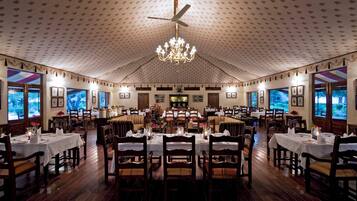 A large dining hall with multiple tables, chandeliers, and framed pictures on the walls.