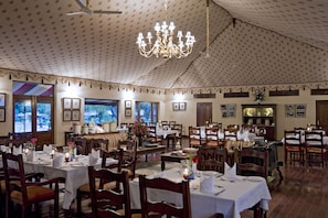 A dining area with multiple tables set for guests, a chandelier, and framed pictures on the walls. - Samode Bagh (Chomu)