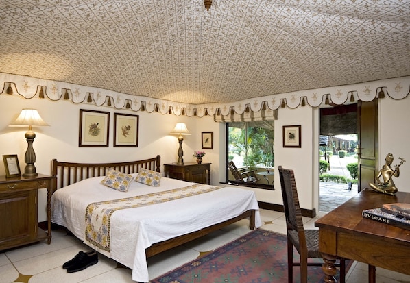 A bedroom with a large bed, two bedside tables with lamps, a wooden chair, a desk with a statue, and framed pictures on the wall. - Samode Bagh (Chomu)
