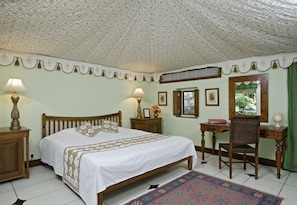 A bedroom with a bed, two bedside tables with lamps, a desk with a chair, a mirror, and a window with a view of greenery. - Samode Bagh (Chomu)