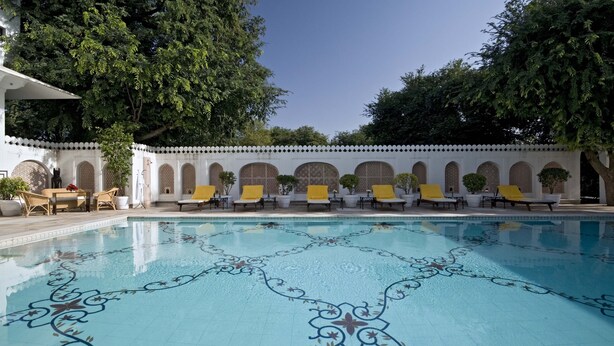 Ein Pool mit geometrischem Fliesenmuster, umgeben von weißen Wänden mit Rundbogenfenstern und gelben Liegestühlen.