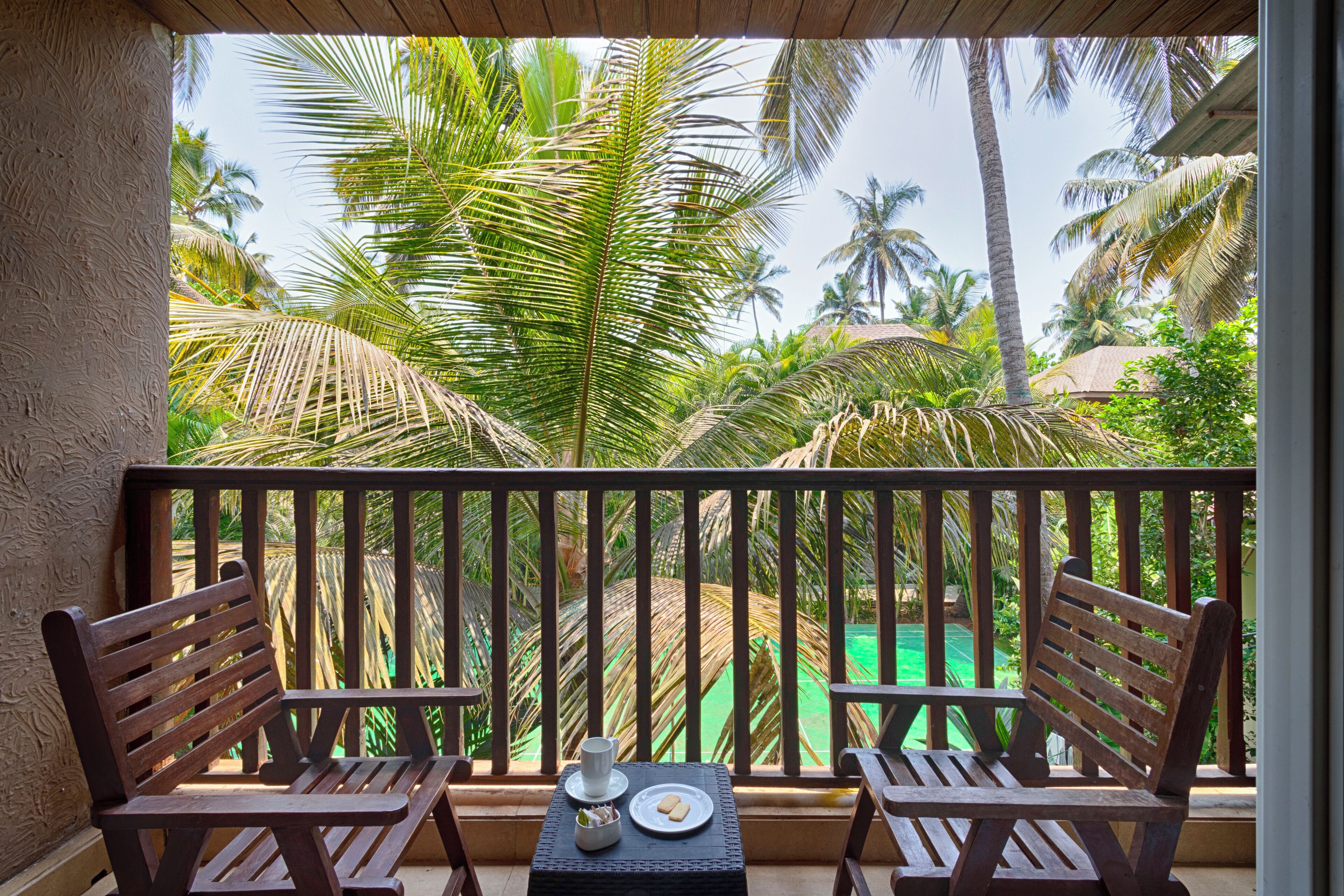 A balcony with two wooden chairs, a small table, and a view of a pool and palm trees.