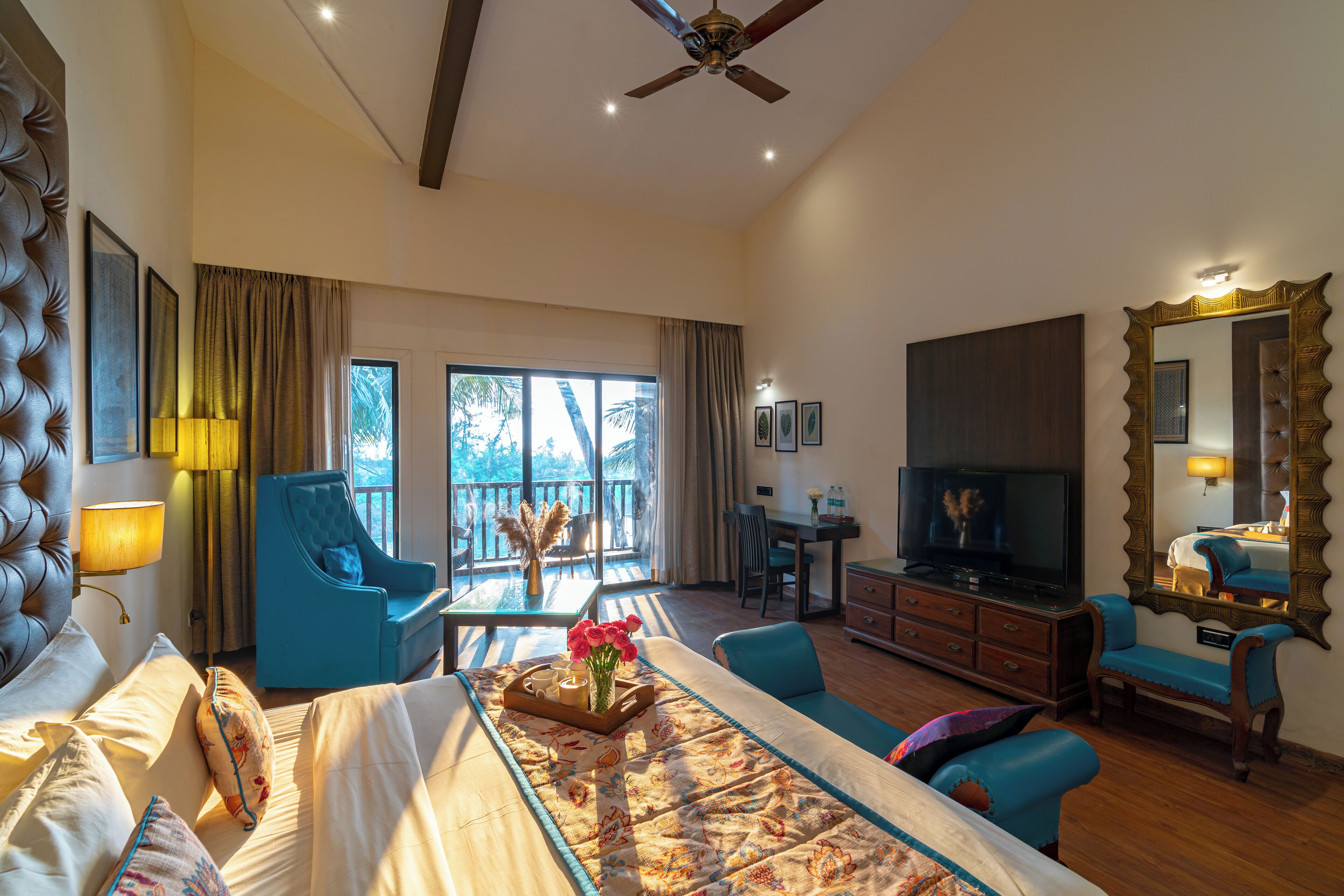 A spacious hotel room with a large bed, a sitting area with a sofa and armchairs, a dining table, a TV, and a balcony with a view.