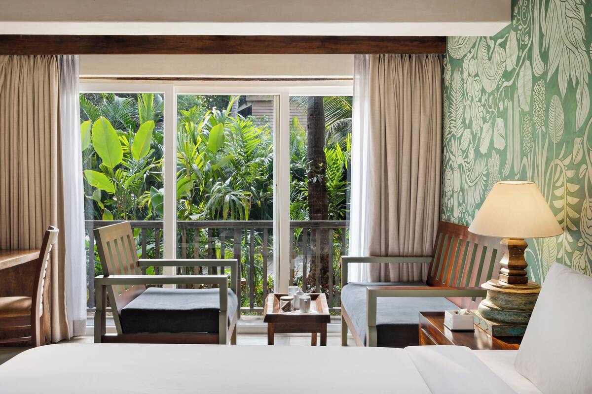 Quarto de hotel com uma janela grande, uma cama, duas cadeiras, uma mesinha e um abajur.