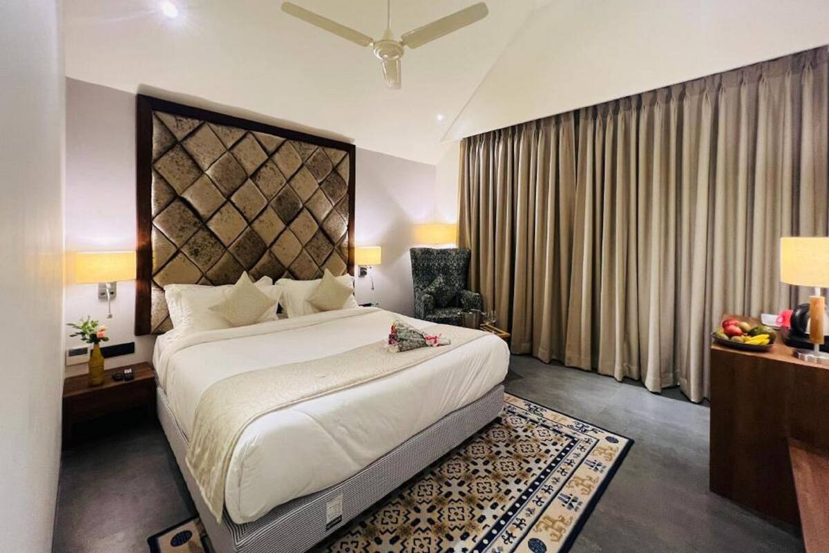 Quarto de hotel com uma cama grande, um ventilador de teto, um tapete estampado e vista para o exterior através de cortinas.