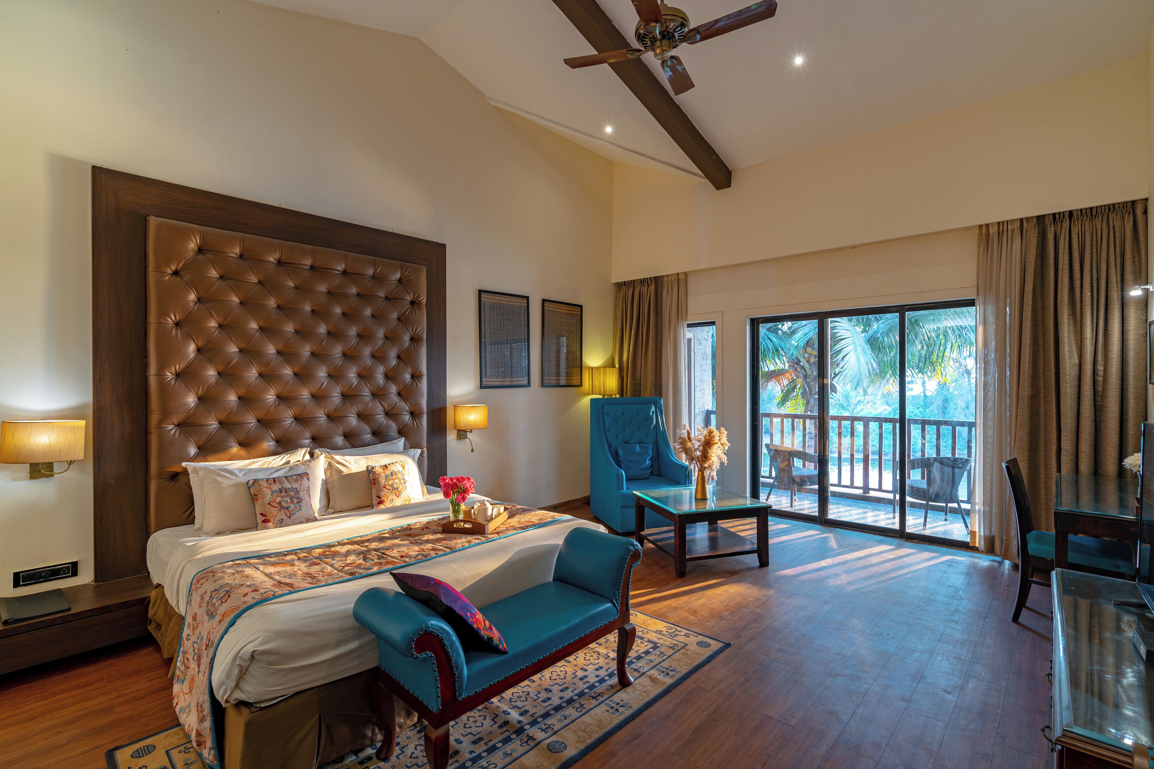 A spacious bedroom with a large bed, a sitting area with a sofa and armchairs, a dining table, and a balcony with a view of palm trees.