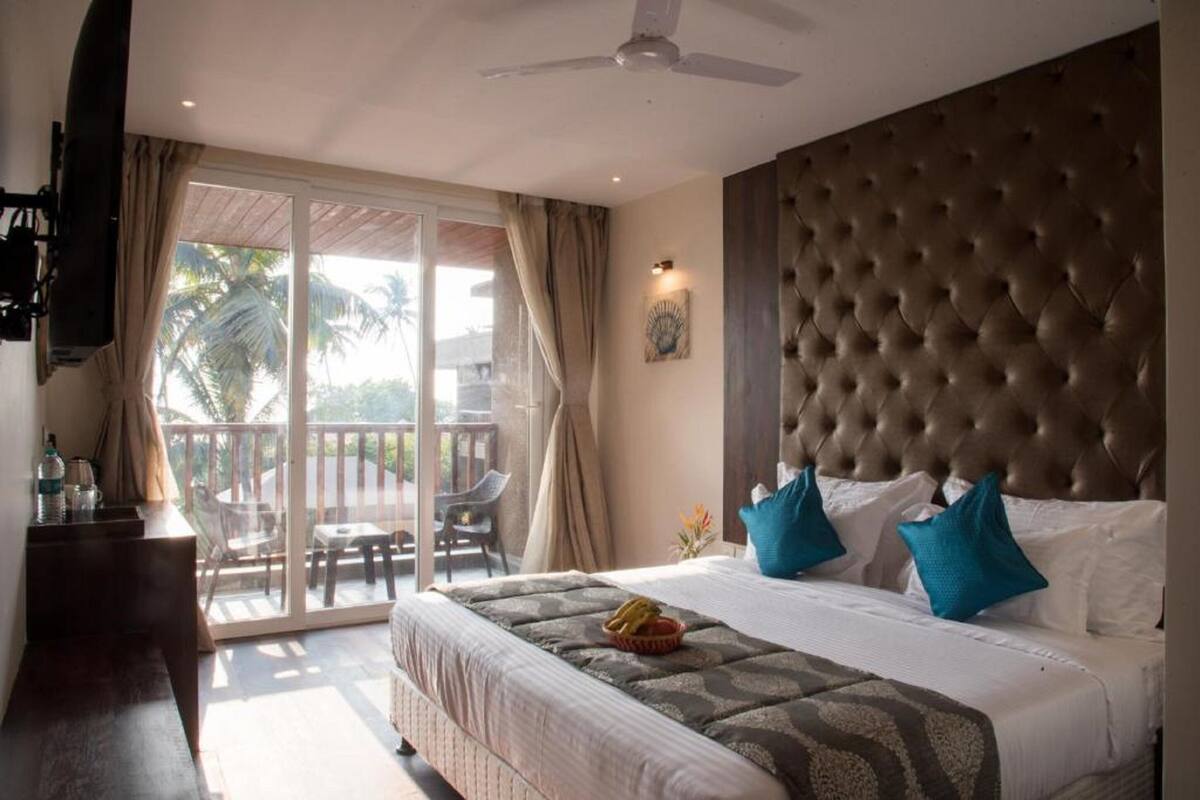 Quarto de hotel com uma cama grande, ventilador de teto, varanda com vista para palmeiras e uma pequena mesa de trabalho com uma televisão.