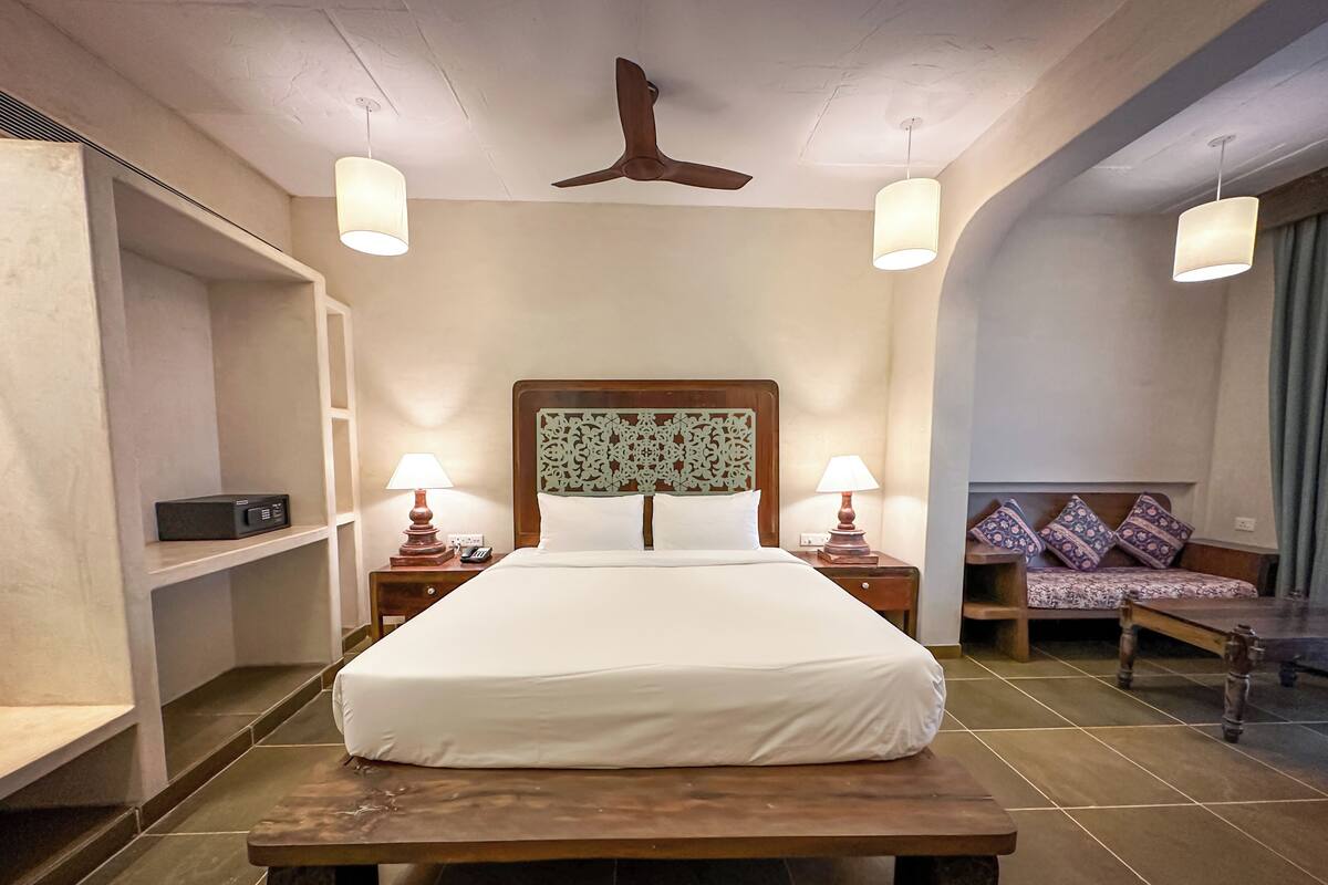 Quarto de hotel com uma cama grande, duas mesas de cabeceira com luminárias, uma cabeceira de madeira, um sofá com almofadas estampadas e um ventilador de teto.