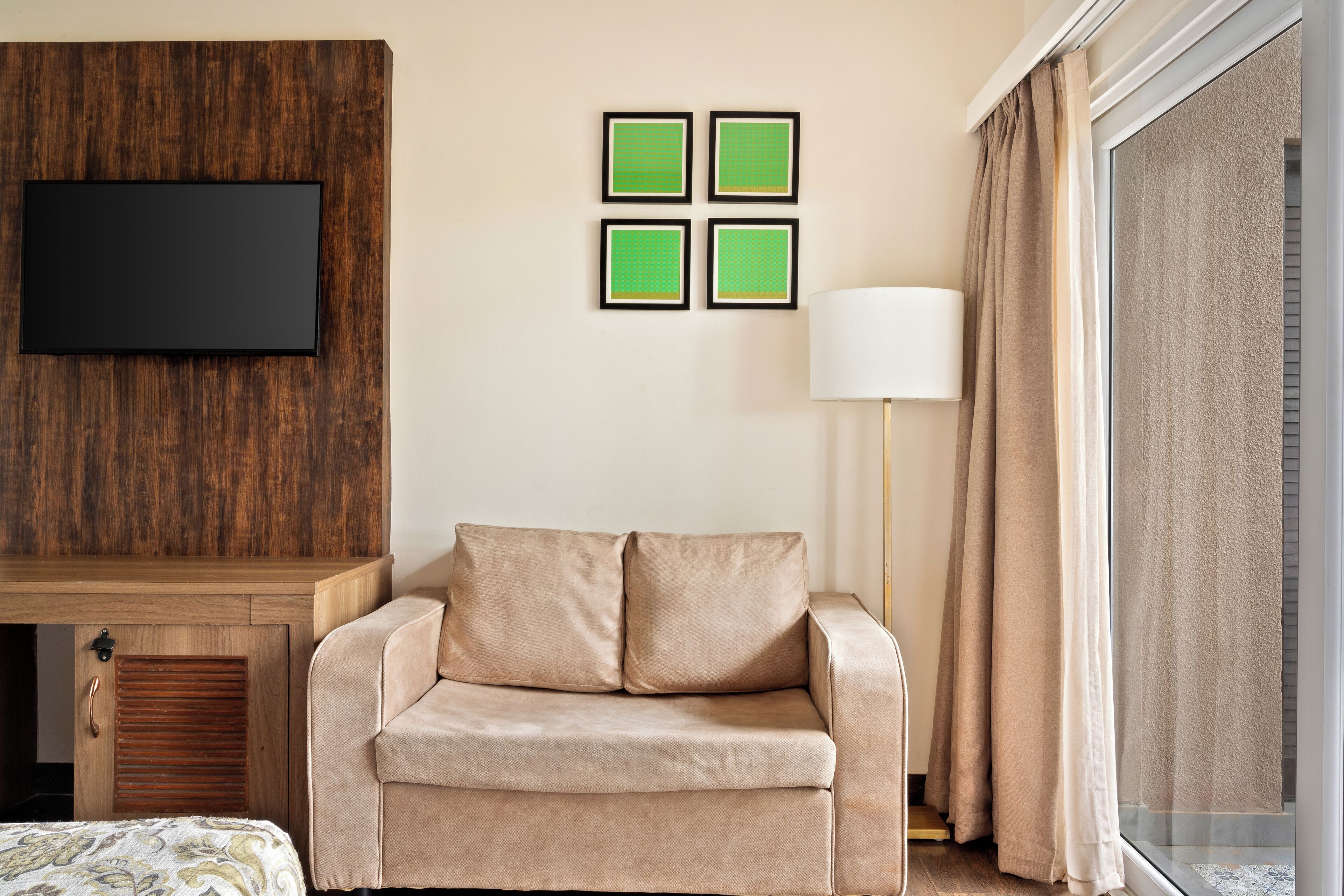 A hotel room with a wooden headboard, a beige sofa, a flat-screen TV mounted on the wall, and a large window with curtains.