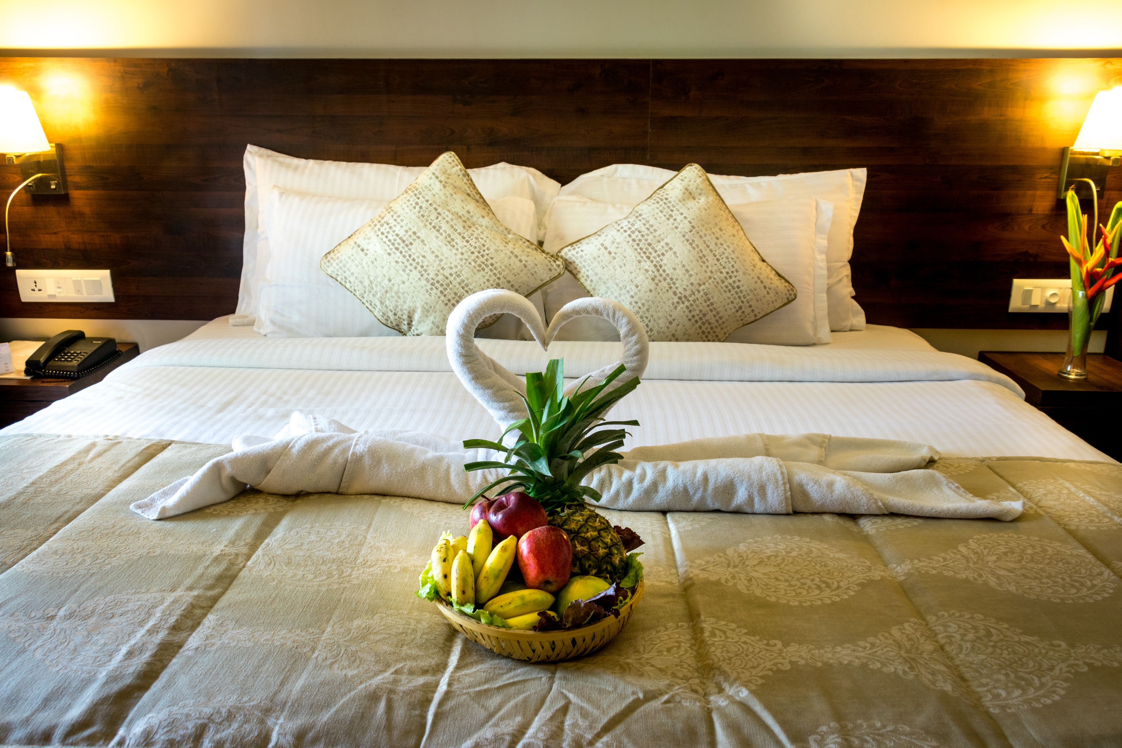 A neatly made bed with white linens, a heart-shaped towel arrangement, and a fruit basket on the bed.