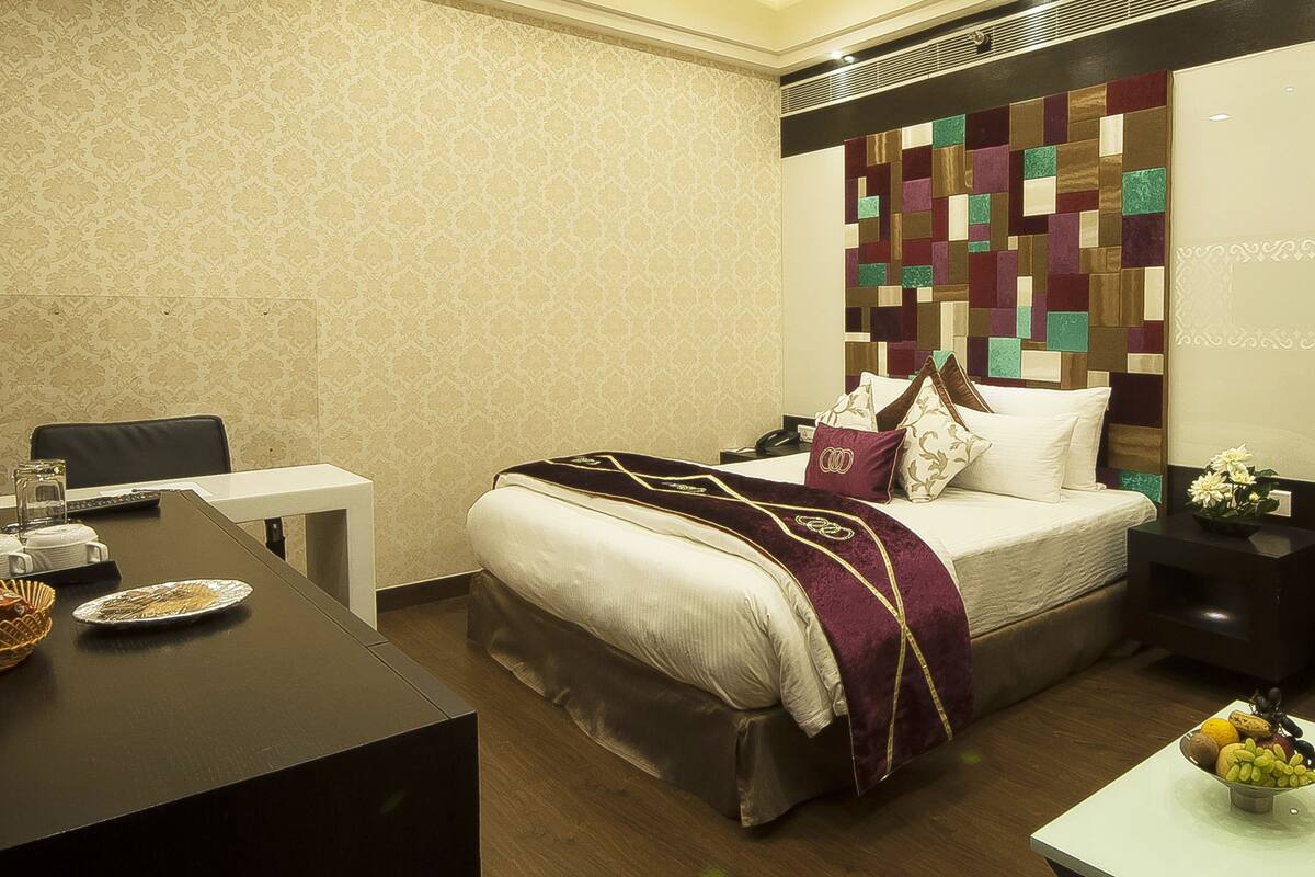 Habitación de hotel con una cama bien tendida, una mesita con frutero, un escritorio con cesta y una silla.