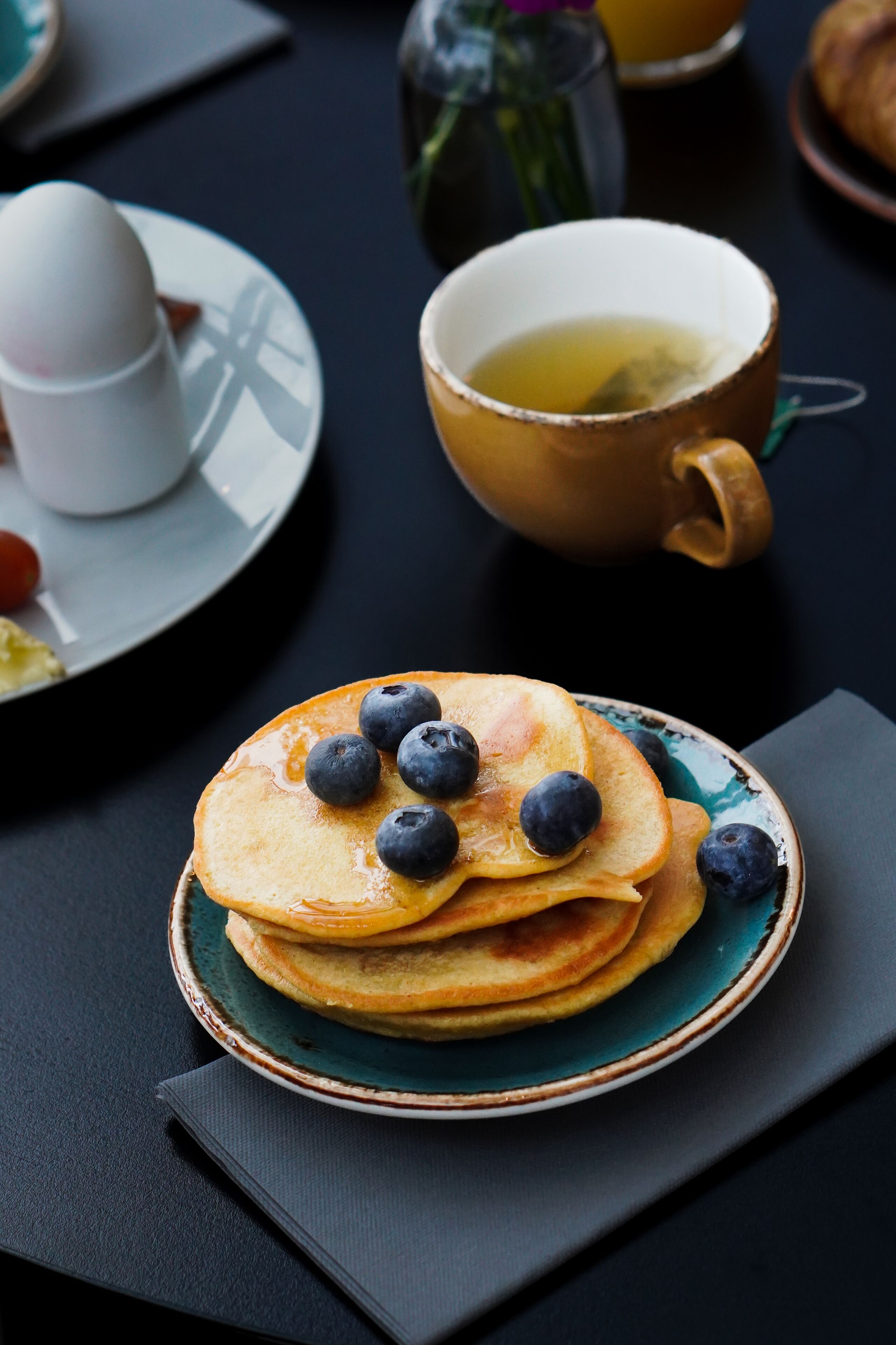 A stack of pancakes with blueberries on a plate, a cup of tea, and a croissant on a table.