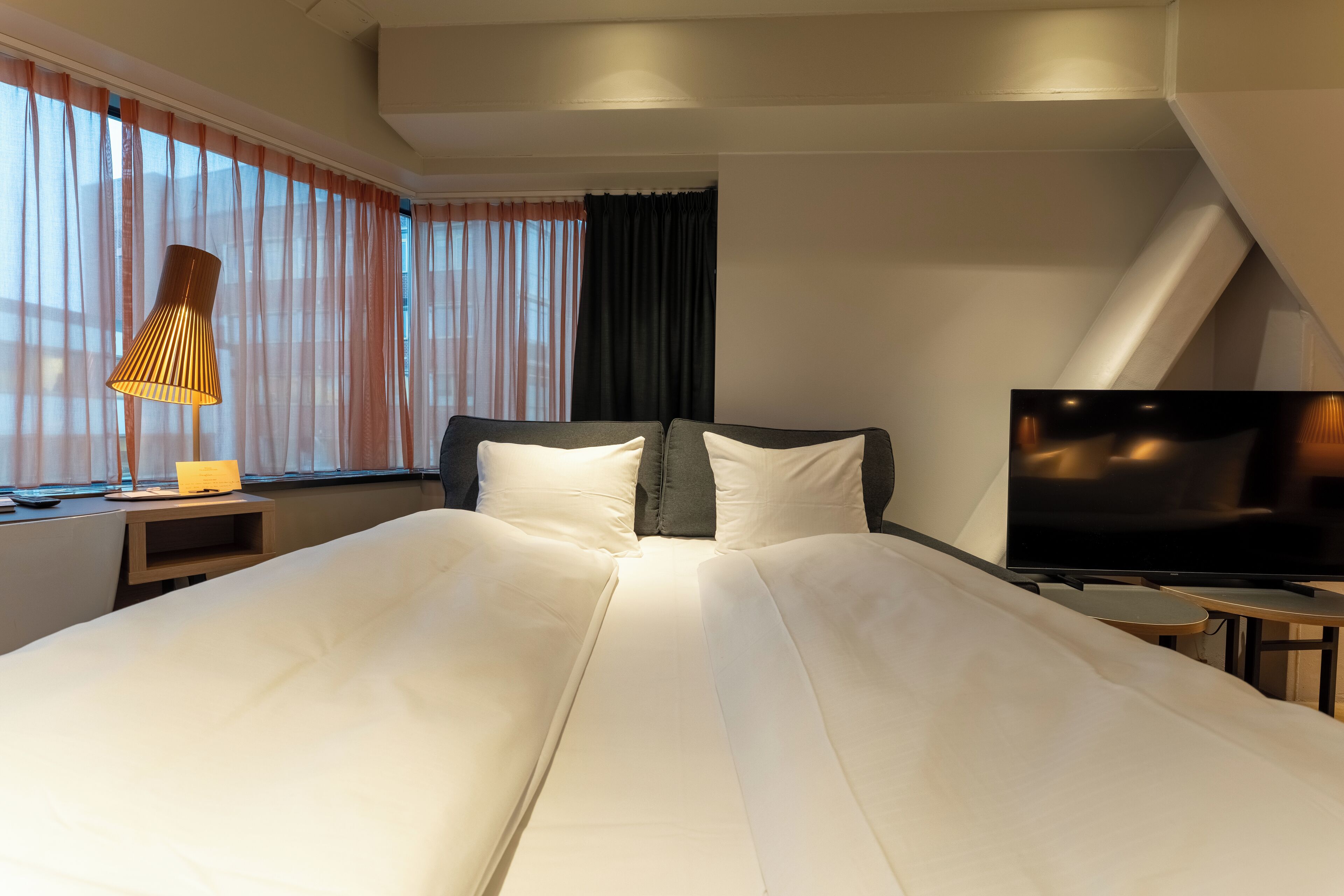 Habitación de hotel con una cama grande, televisión, lámpara y ventana con cortinas.