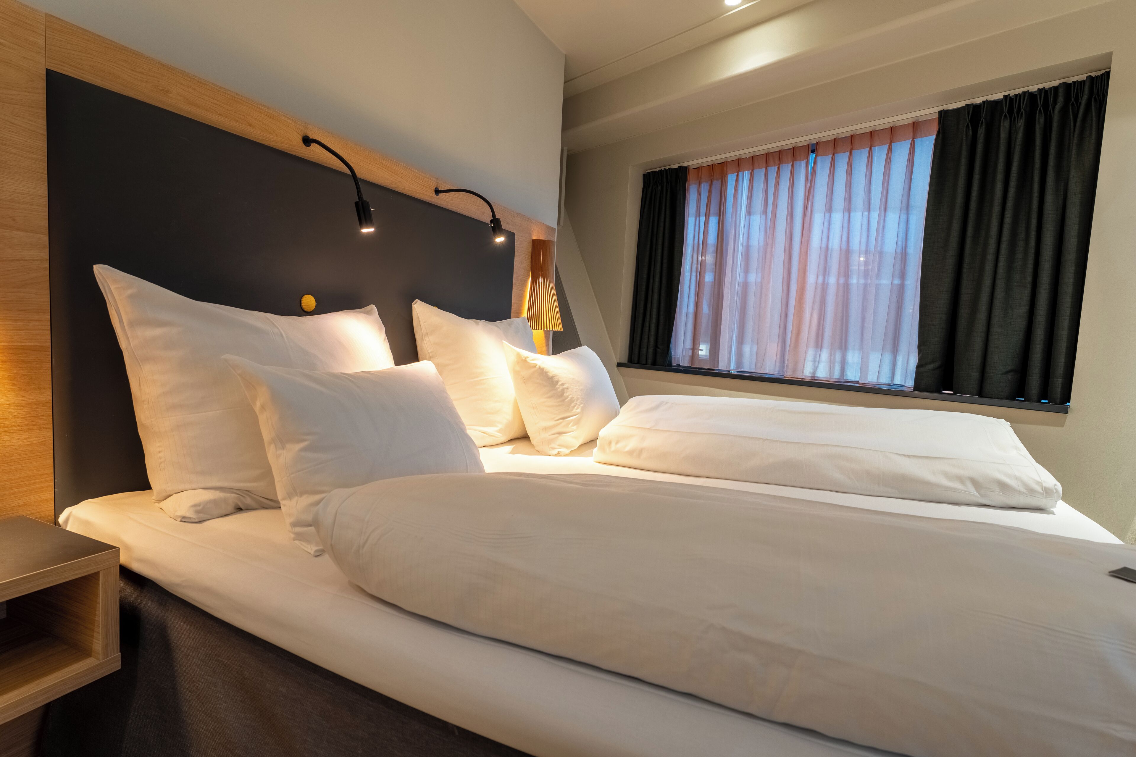 Une chambre d’hôtel avec un grand lit, une table de chevet, une lampe et une fenêtre avec des rideaux.