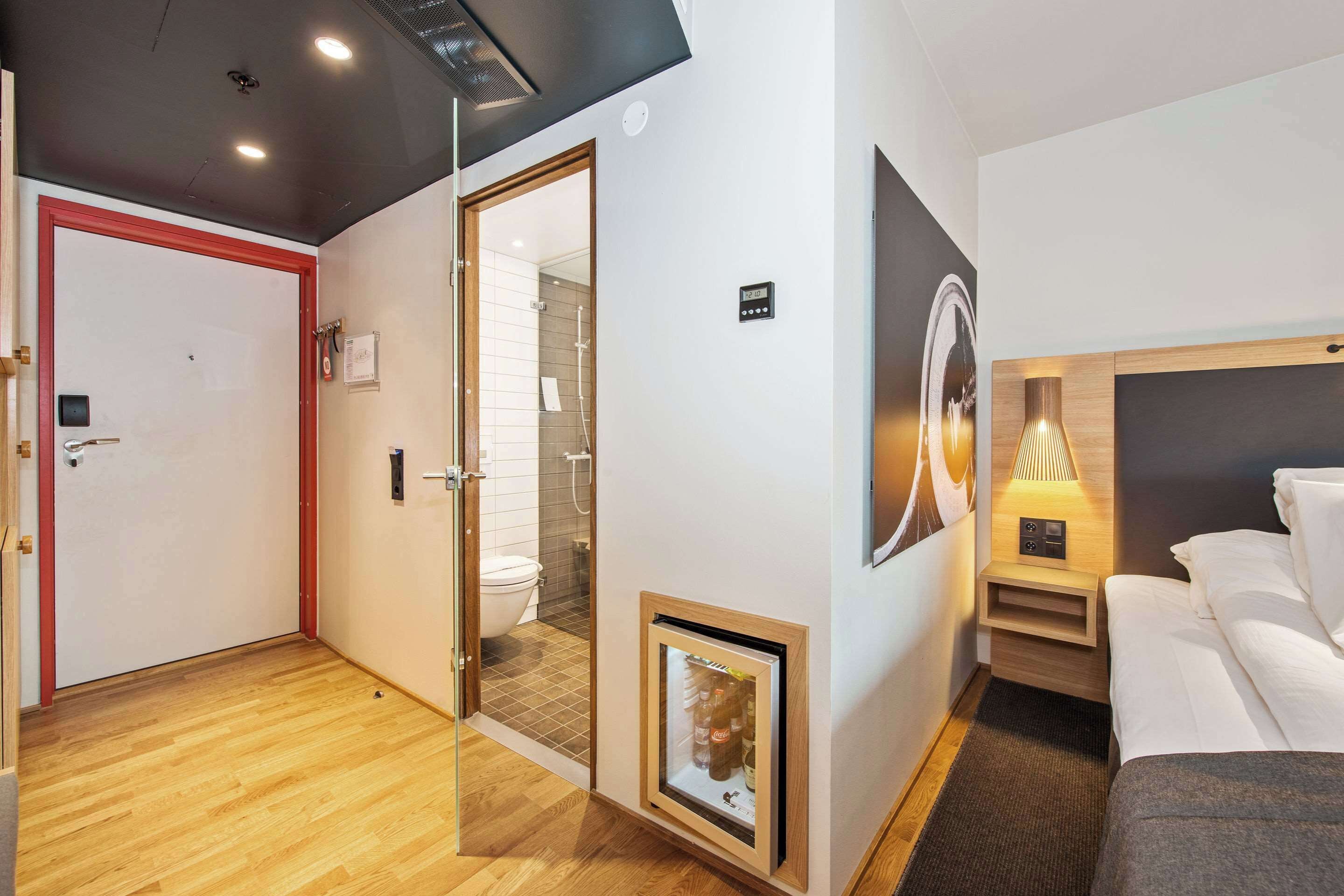 Una habitación de hotel con suelo de madera, una cama con ropa de cama blanca, una mesita de noche con lámpara, un cuadro enmarcado en la pared y un baño visible a través de una puerta abierta.