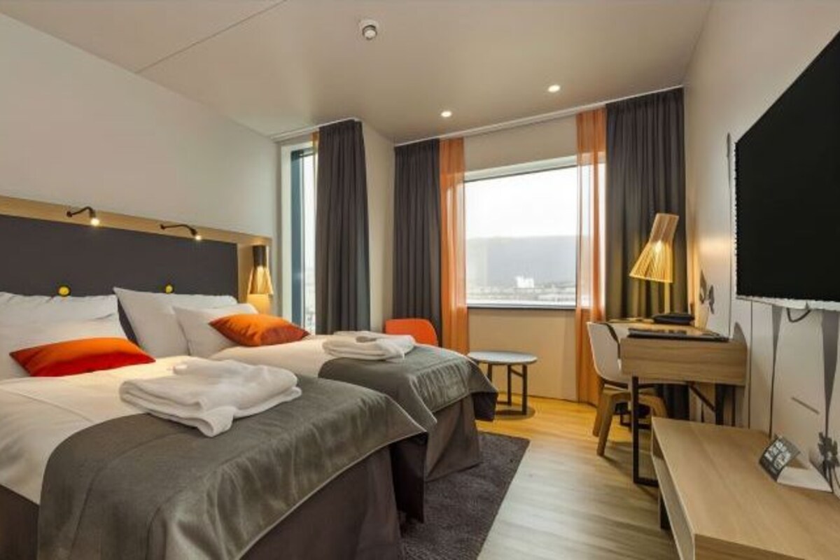 Una habitación de hotel con dos camas, un escritorio, una silla y un ventanal con cortinas.