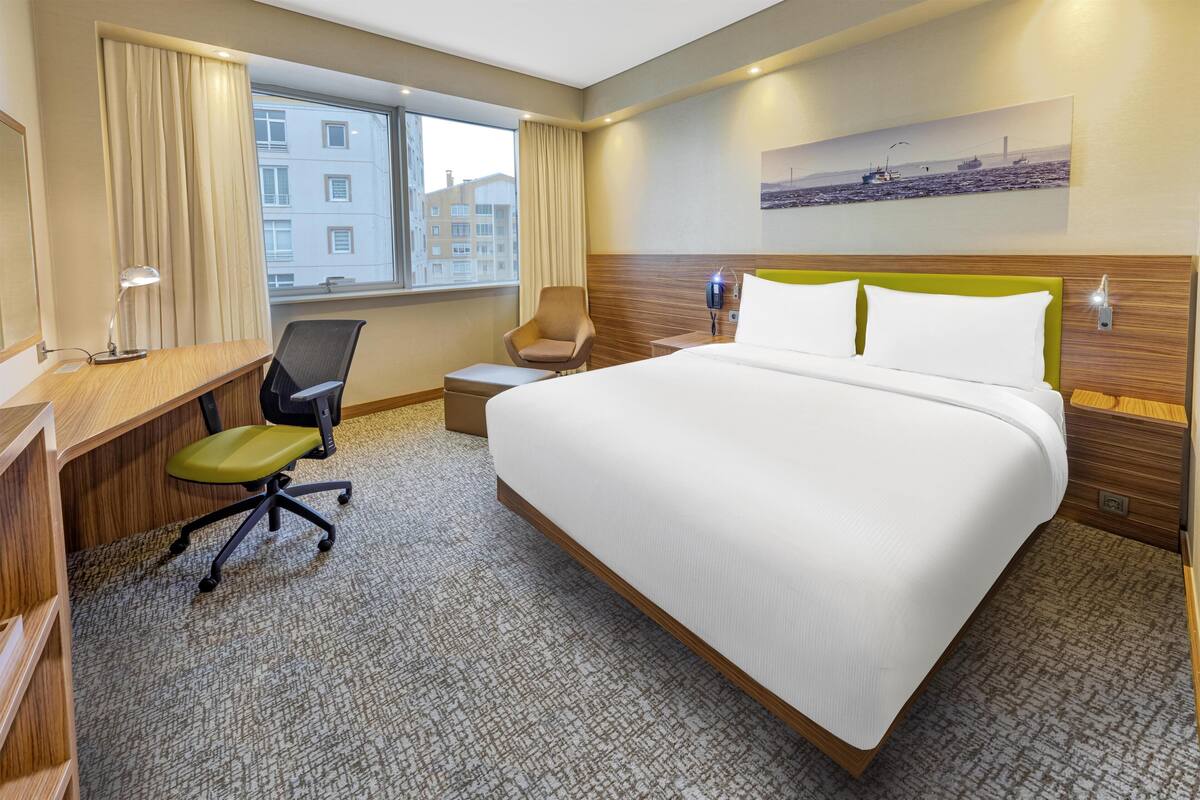 Una habitació d'hotel amb un llit gran, un escriptori amb una cadira, una taula petita, una finestra amb cortines i un quadre penjat a la paret.